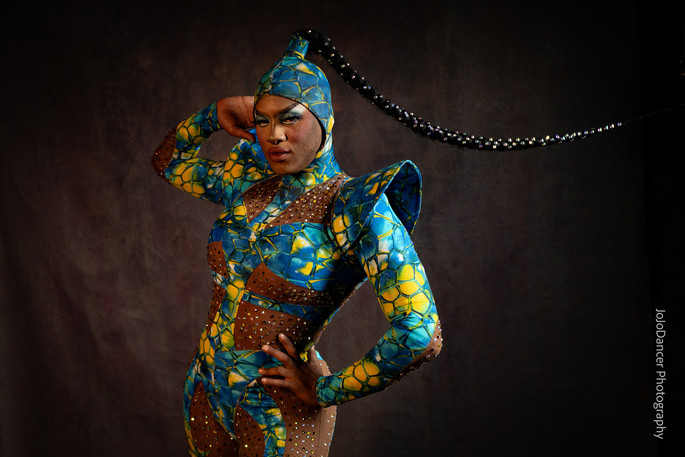 Woman in colorful, patterned costume with headpiece, posing with hand on hip against dark background.