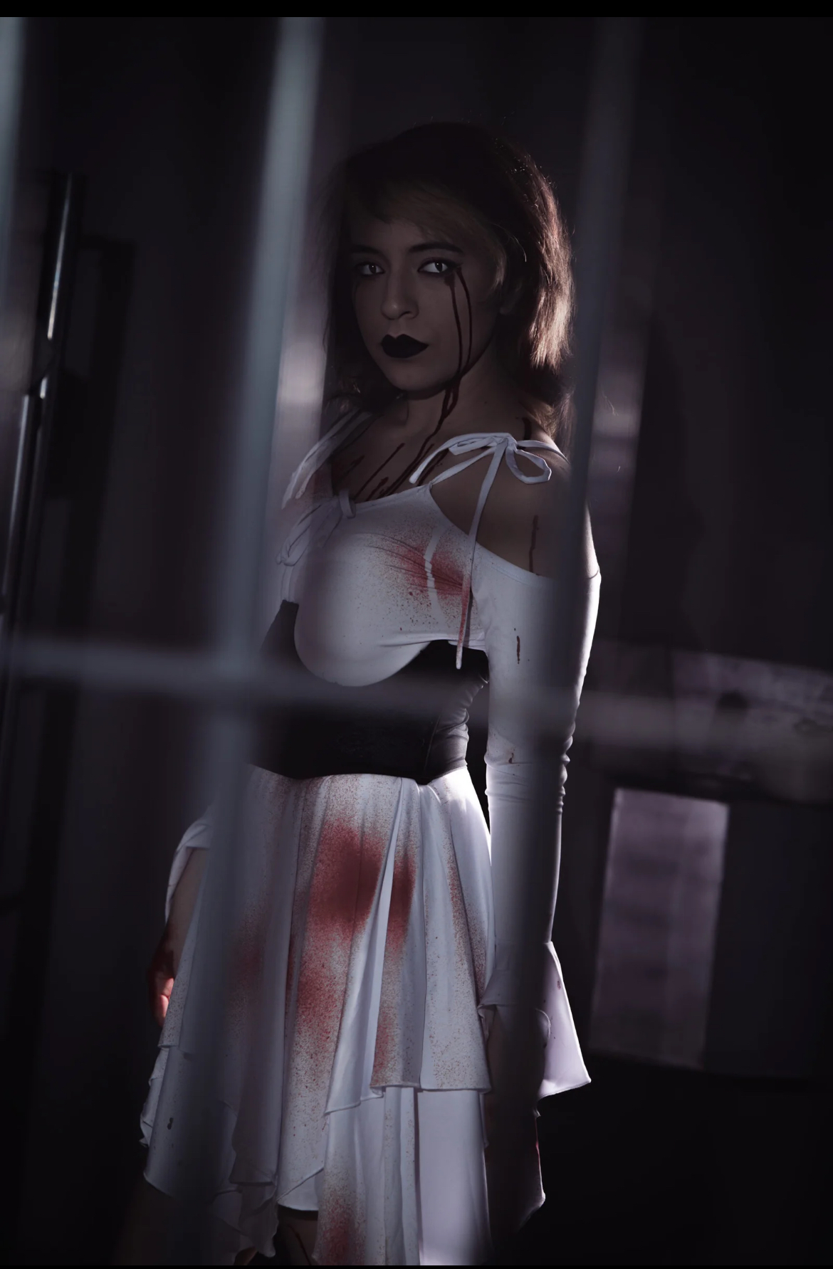 Young woman with dark makeup and hair in a white dress, standing behind bars in a dimly lit setting.