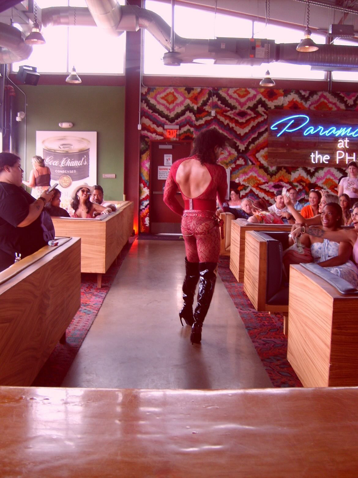Person walking in a colorful restaurant with neon sign and patterned wall, surrounded by seated diners, wooden booths, and vibrant decor.