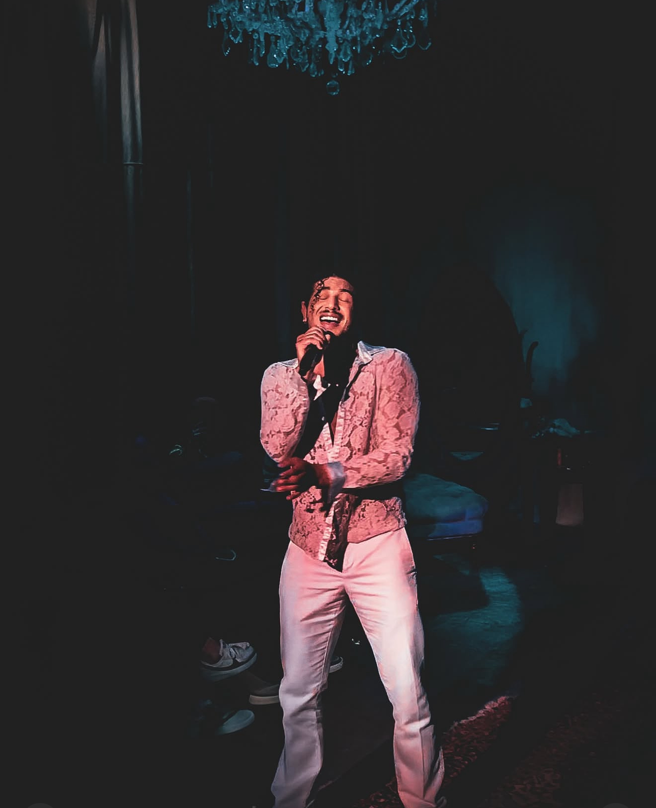 Person singing on stage with microphone, wearing pink jacket and white pants, dark background, blue lighting overhead.