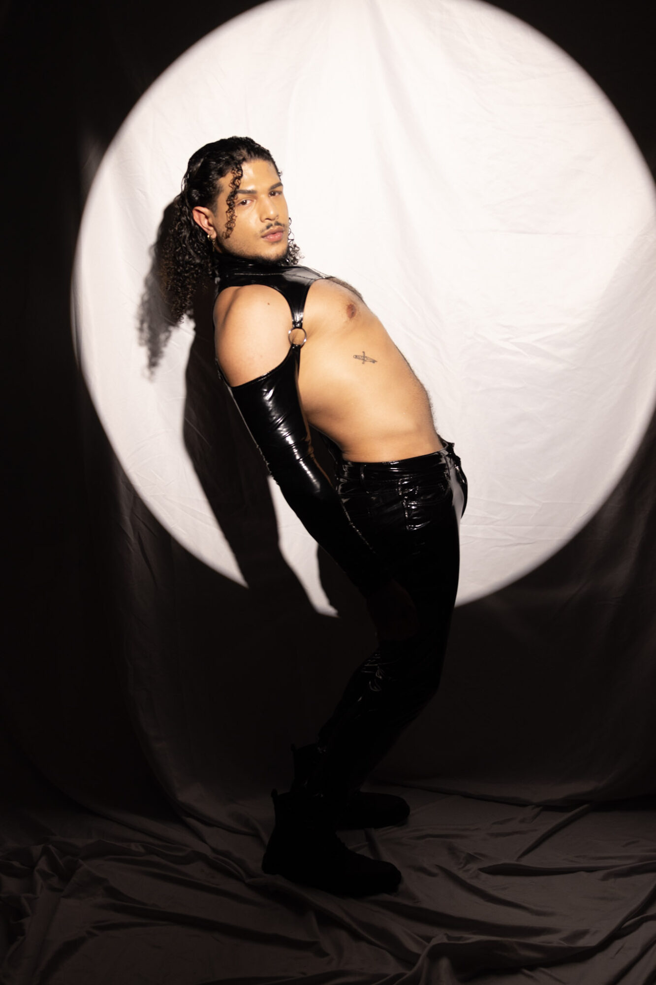 Person with long hair, wearing black gloves and pants, poses against a white circular backdrop, looking to the side.