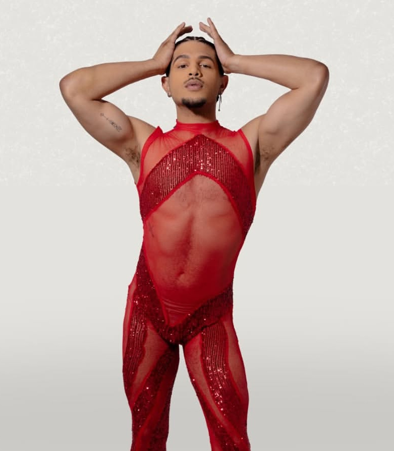 Person in red sheer outfit with arms raised, hands on head, standing against a plain background.
