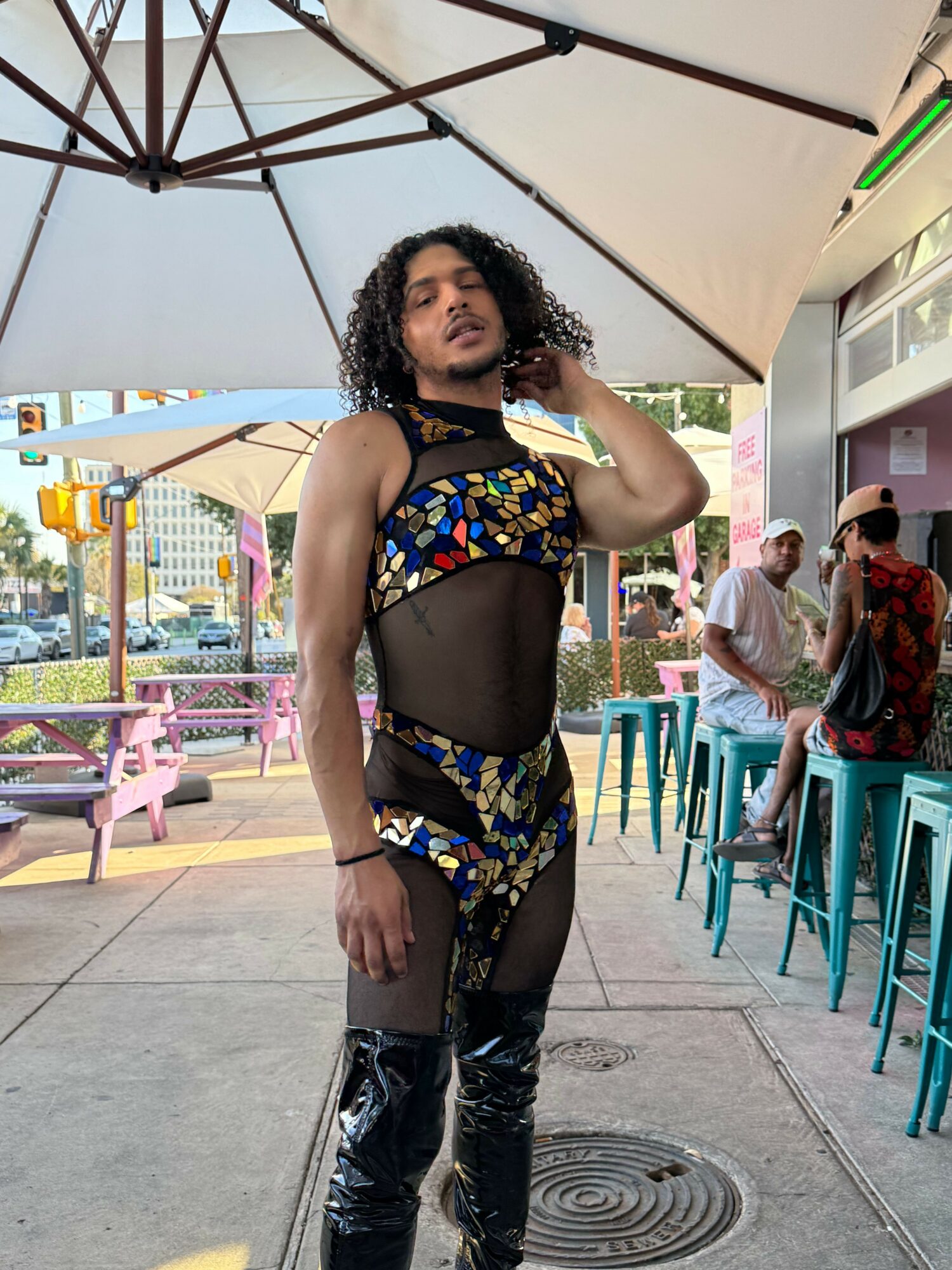 Woman with curly hair in colorful outfit and black tights standing outside near tables and umbrellas.