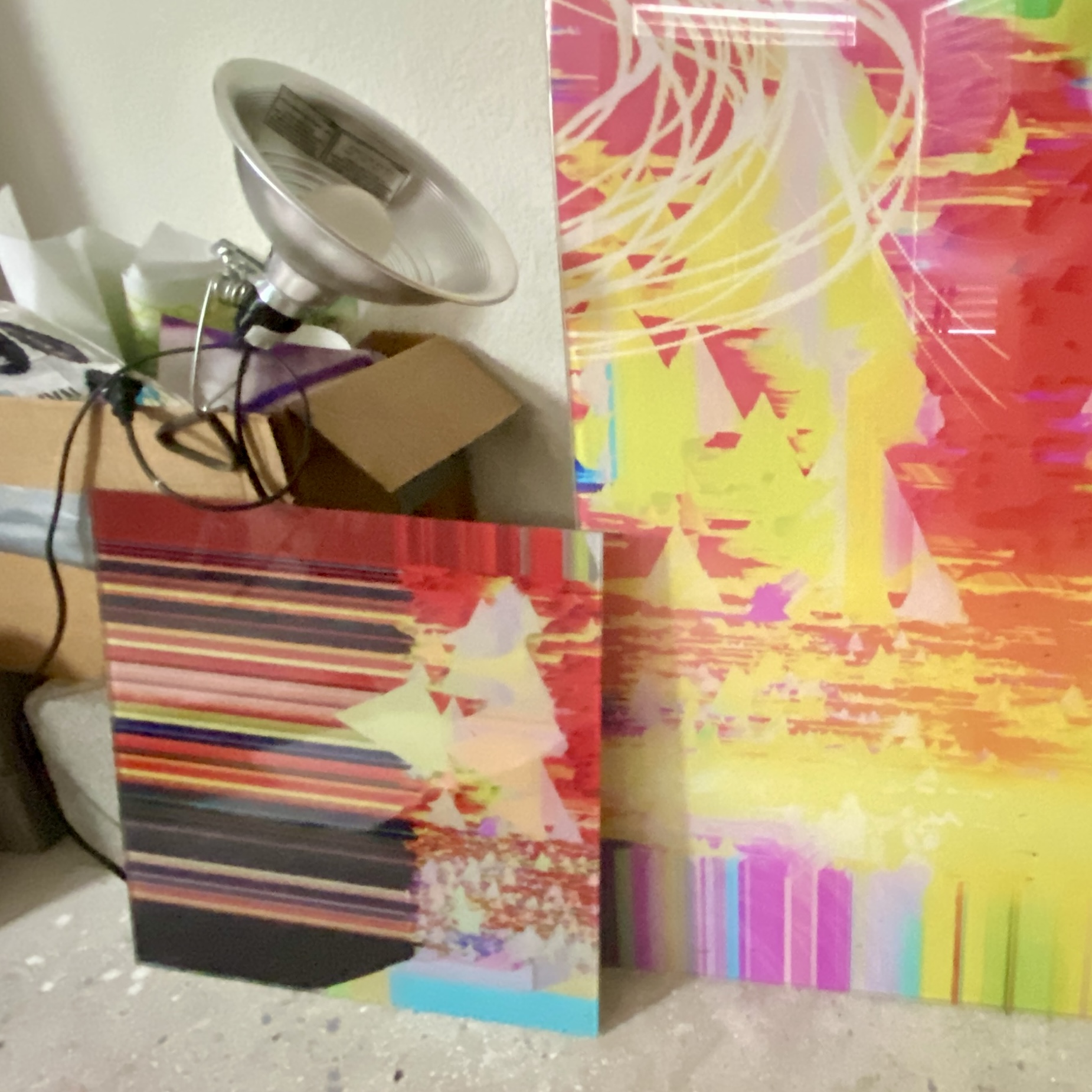 Two colorful abstract paintings leaning against a wall, with a lamp and other objects in the background.