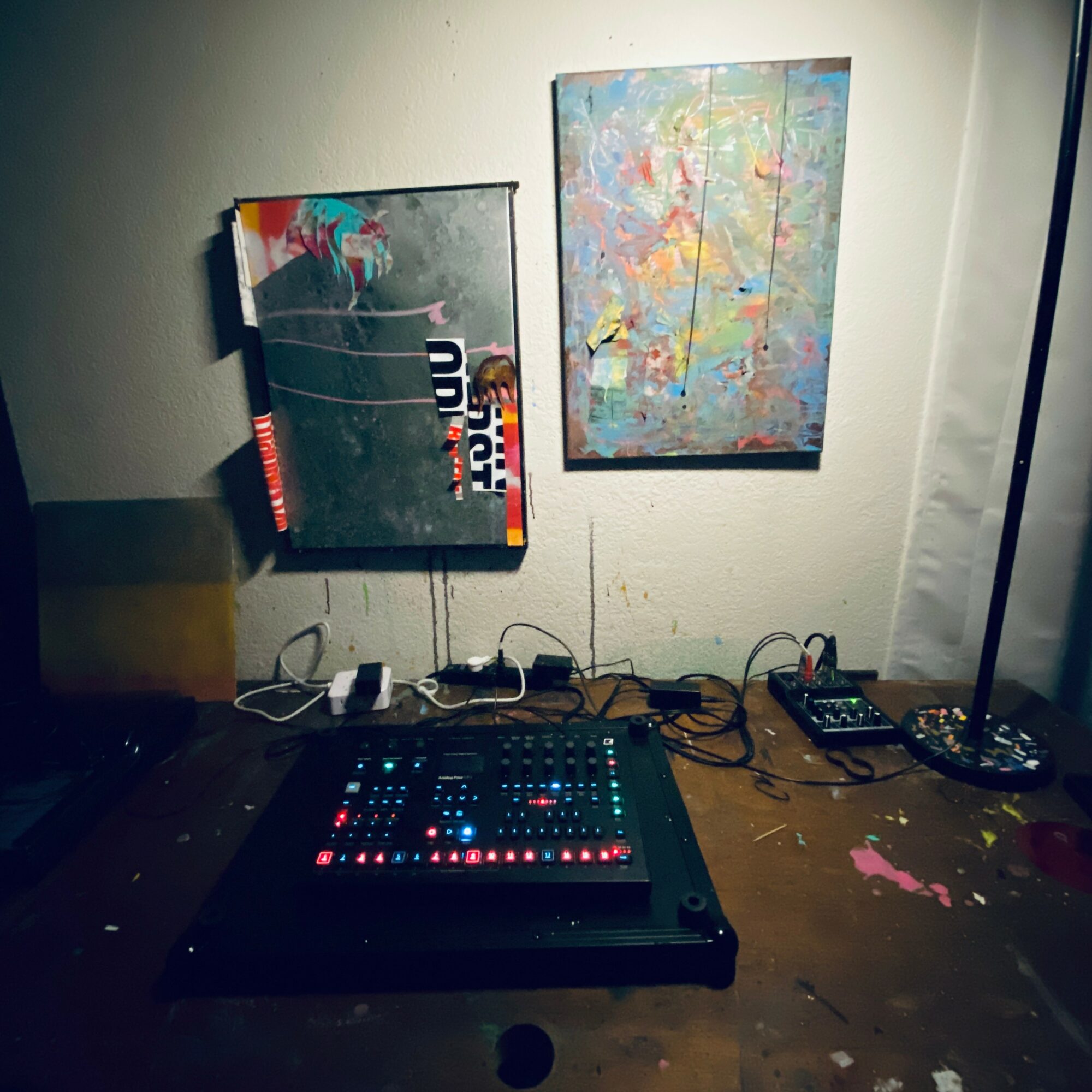 DJ controller on a wooden table with abstract paintings on the wall behind, wires connected to devices, and a lamp on the right.