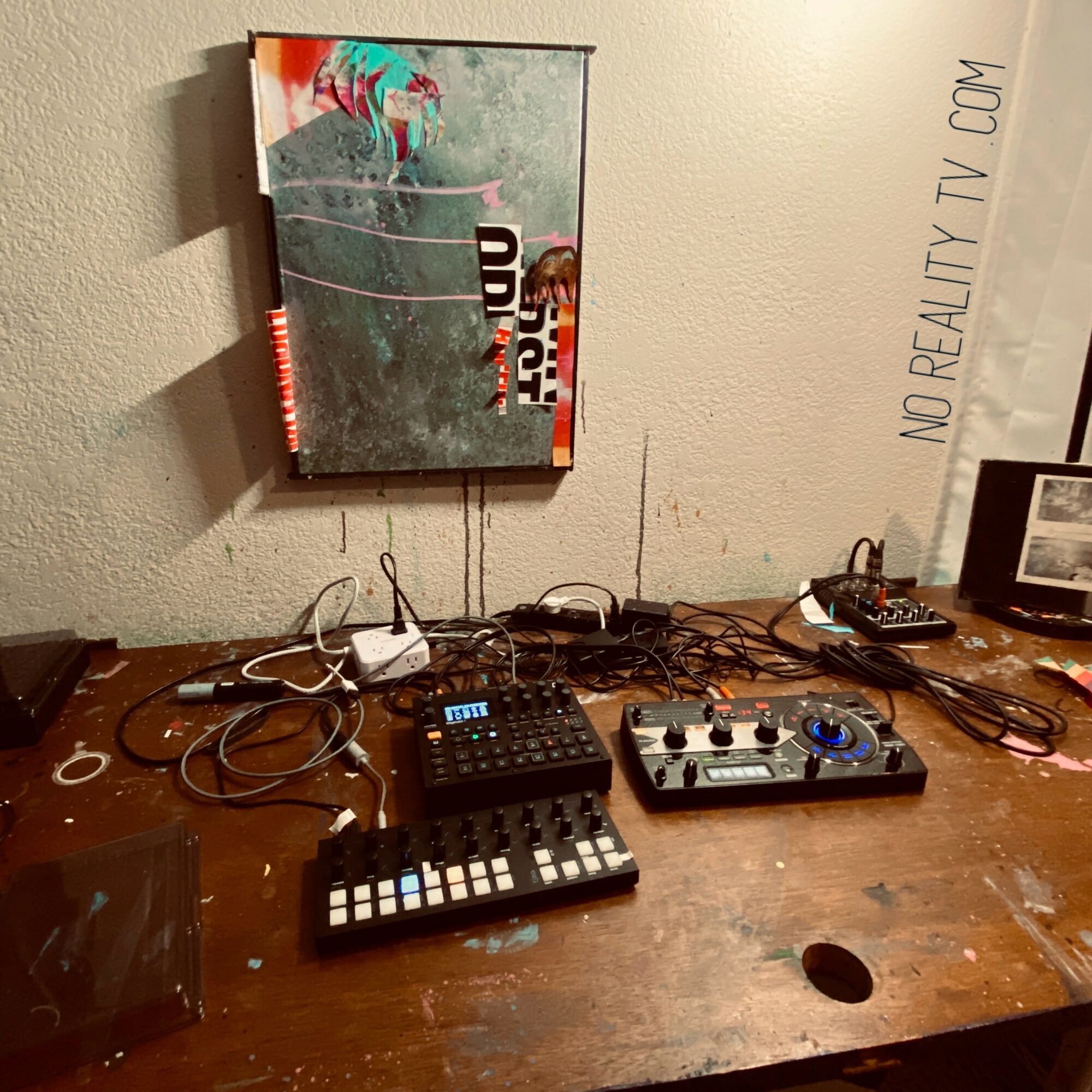 Music equipment on a wooden table with a poster on the wall behind, including a small keyboard, mixer, and cables.