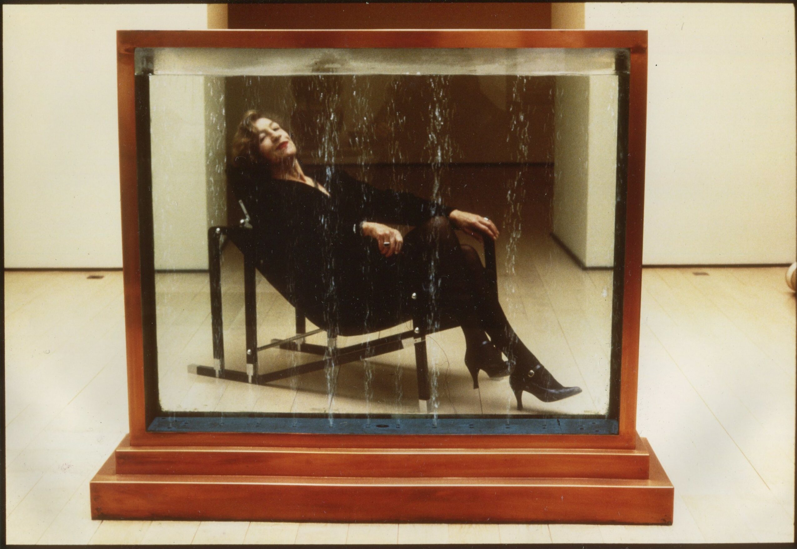 Person sitting on a chair inside a glass display case, reflected in the glass, with a wooden base and background.