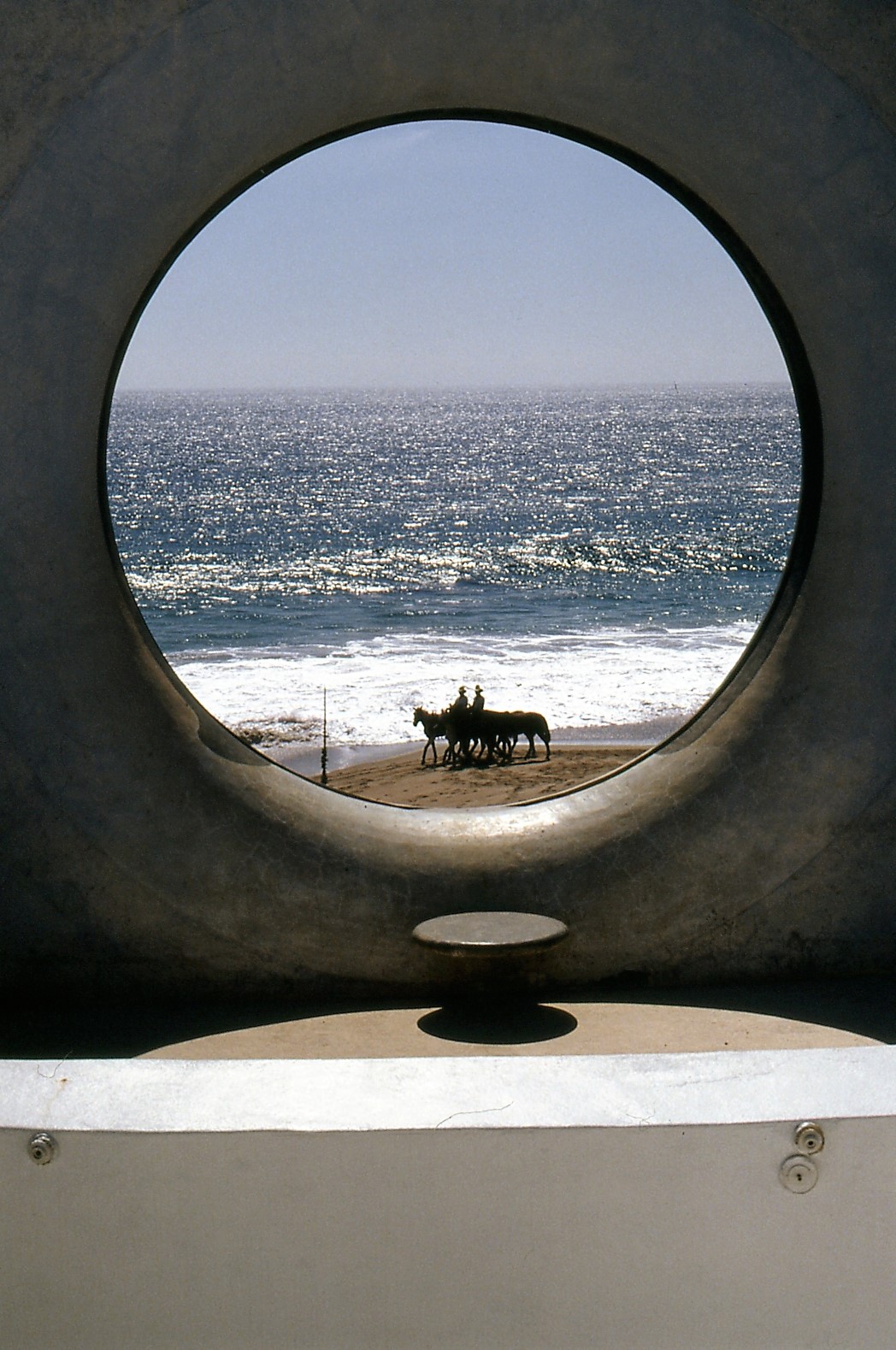 View of the ocean through a circular window, with a horse-drawn carriage on the beach, sky and sea in the background.