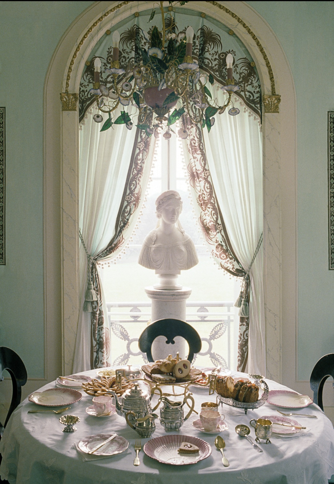 A table set for a meal with a statue of a child on a window ledge behind, decorated with curtains and floral arrangements.