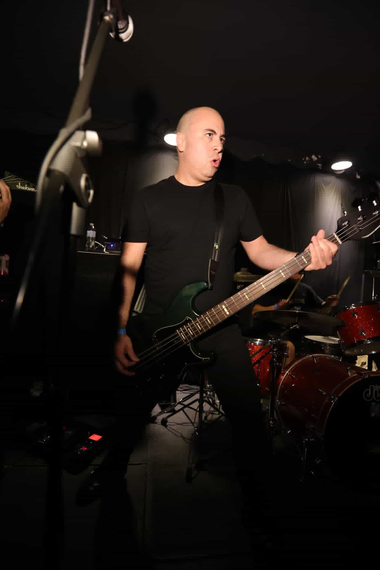 Bald person playing an electric guitar on stage with drums in the background, dark setting, focused expression.