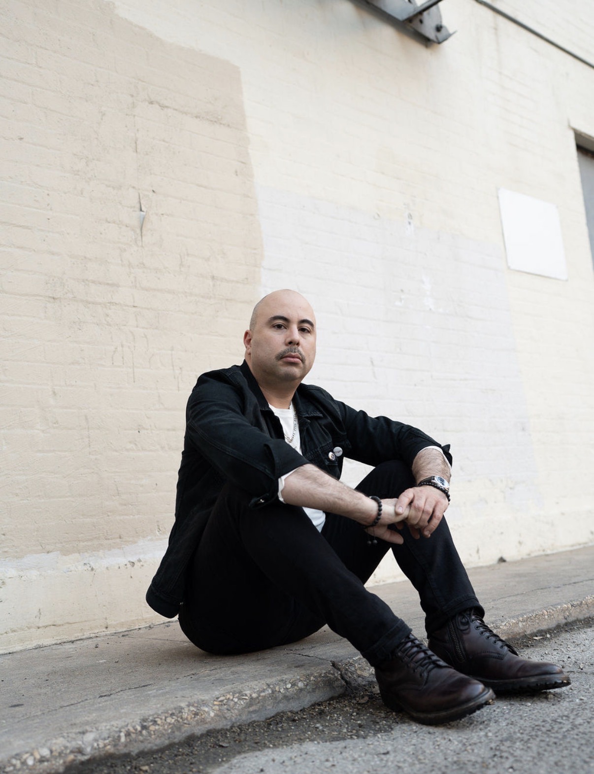 Person sitting on the ground against a beige wall, wearing black clothing and boots, with arms resting on knees.