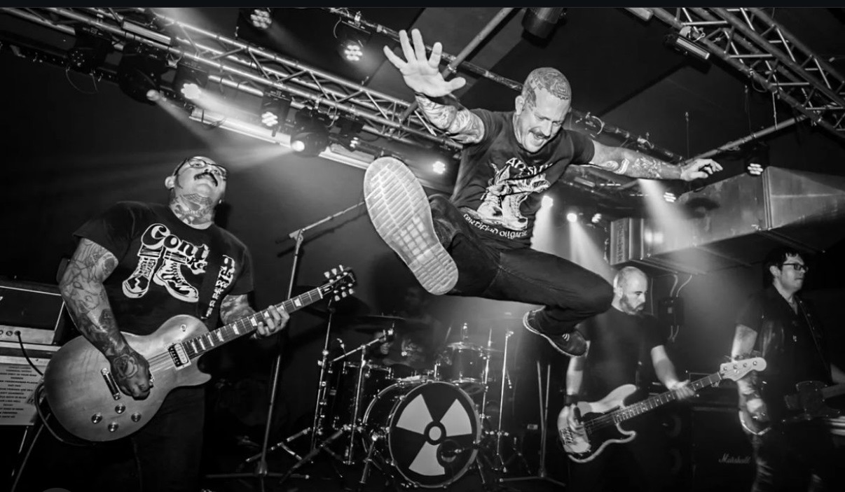 Band performing on stage with guitarist, drummer, and bassist, one member jumping mid-air, black and white photo.