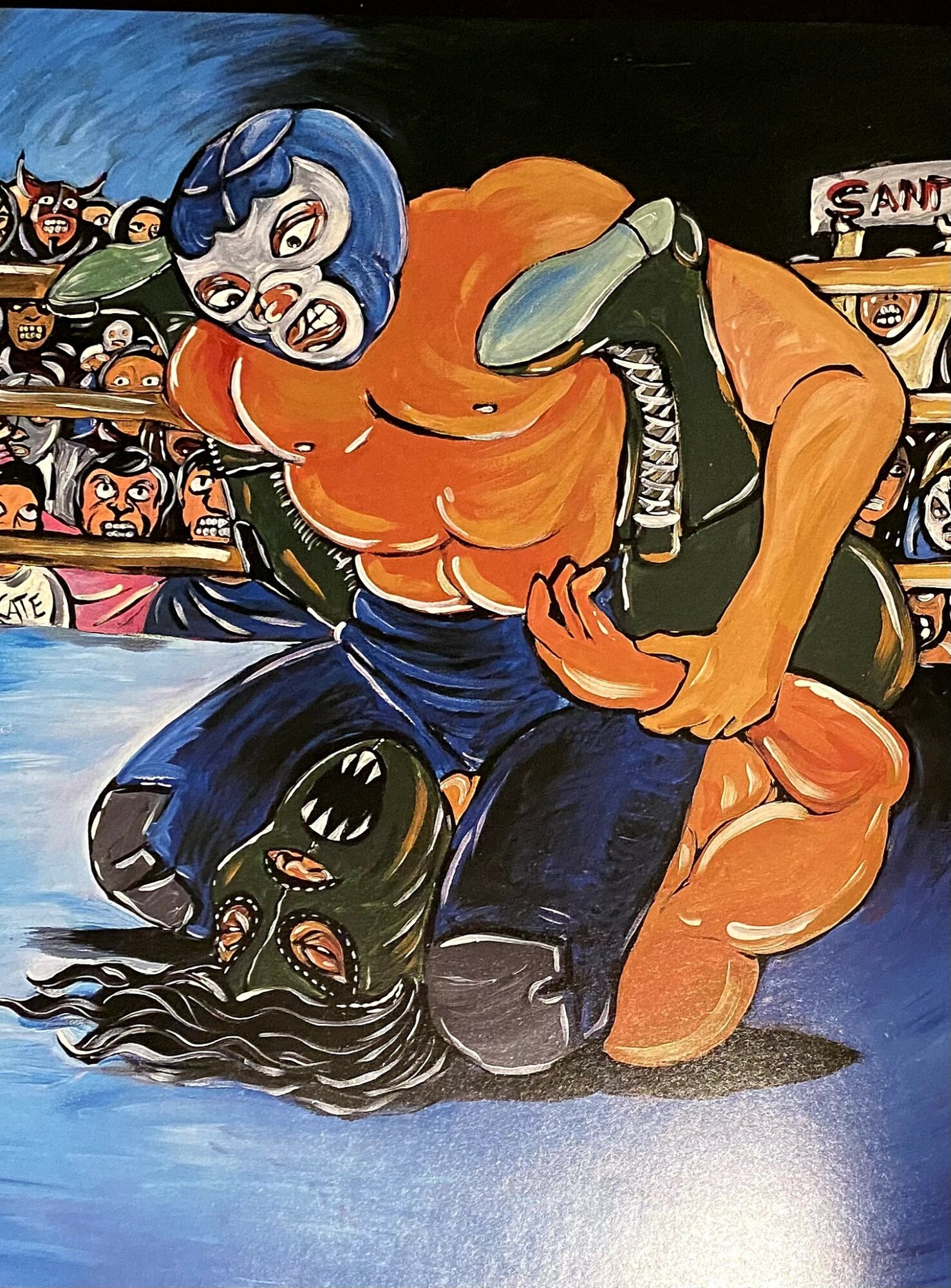 Wrestler in orange and blue mask pins opponent with long black hair in wrestling ring, audience watching in background.