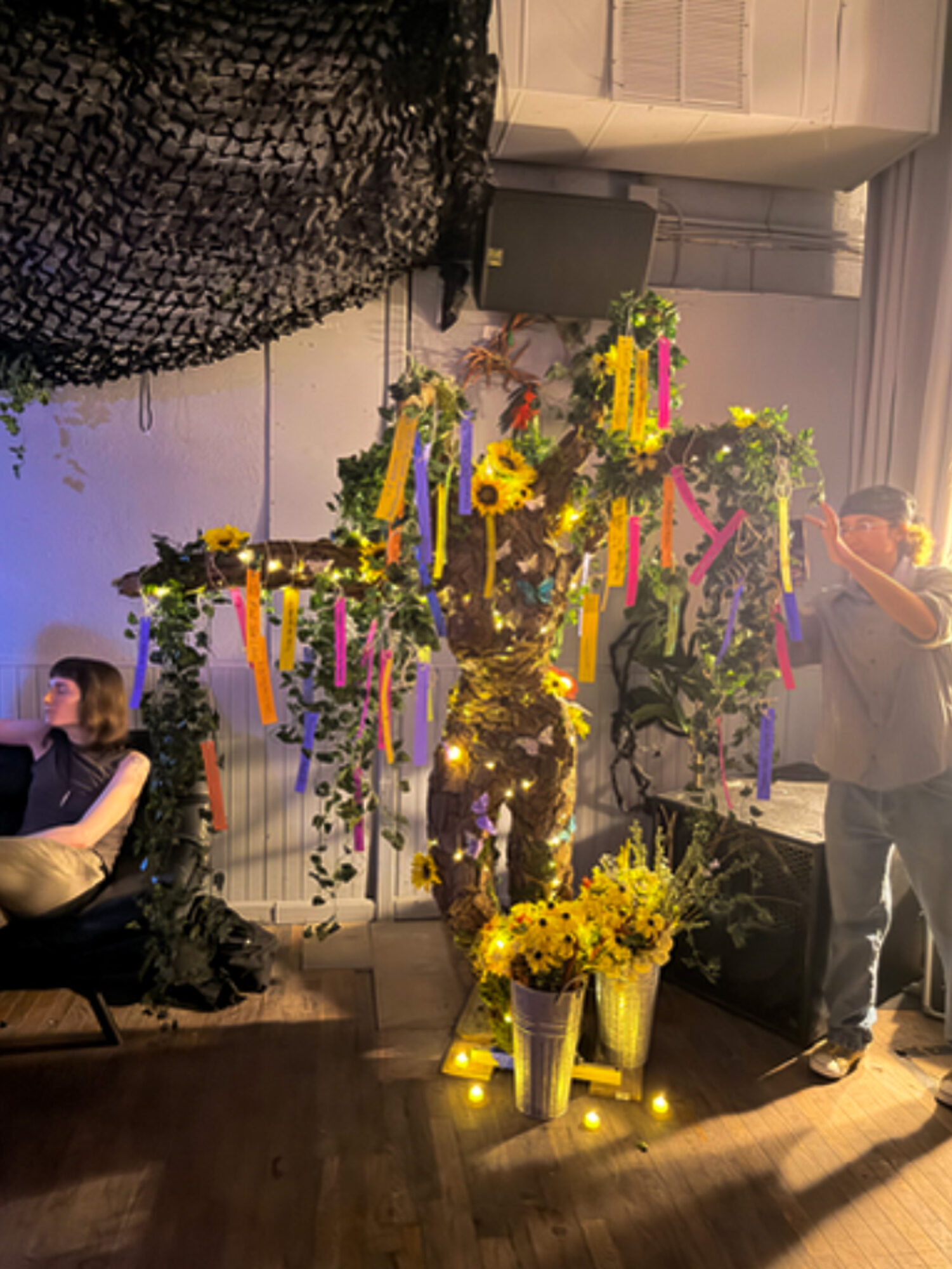 Decorative tree with yellow flowers and hanging ribbons, illuminated by warm lighting, in a room with people and a photographer.