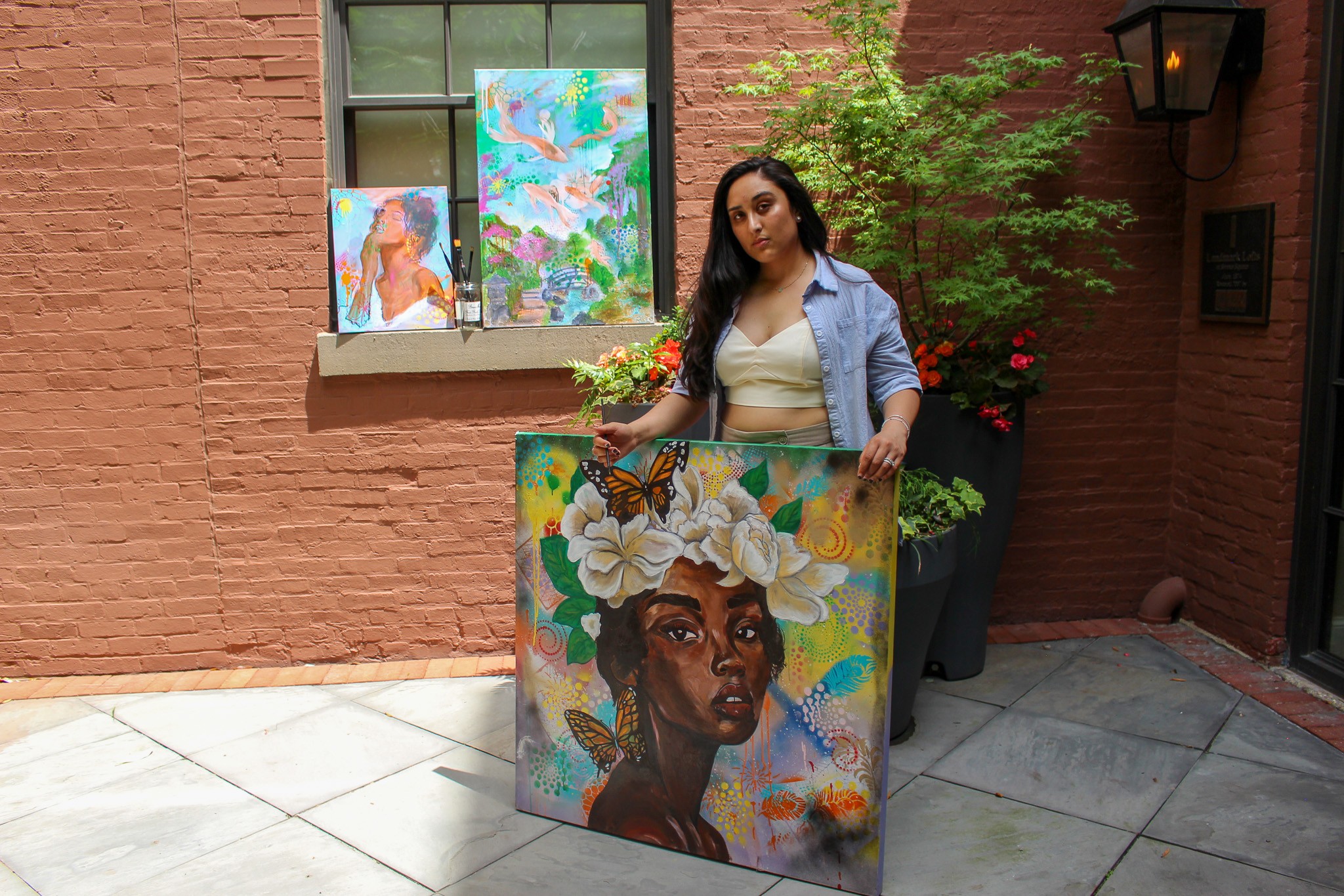 Woman standing outdoors holding a colorful painting of a woman with flowers in her hair, with paintings on the window behind her.
