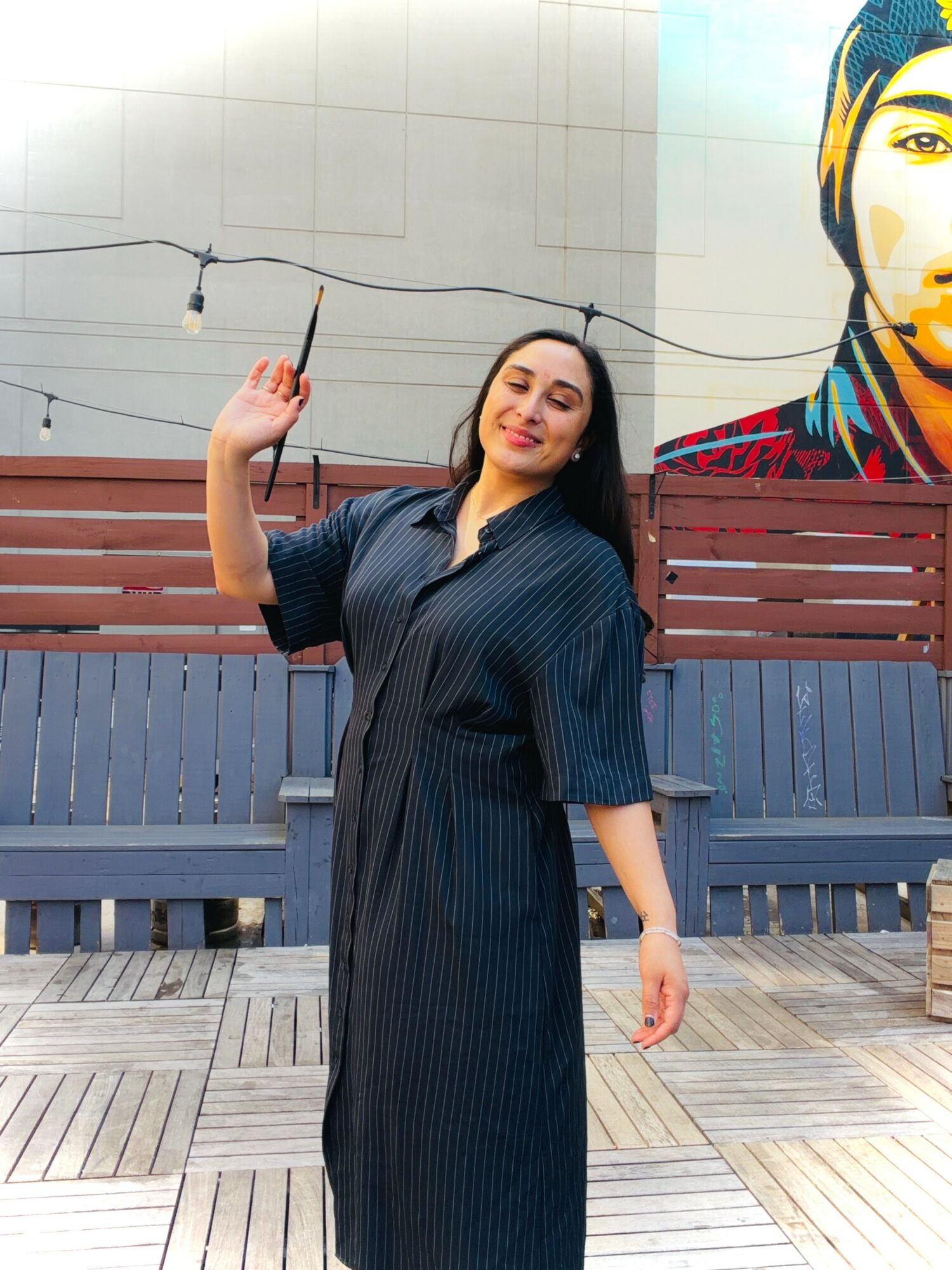 Woman standing outdoors, smiling, wearing a dark striped dress, with a mural of a person's face in the background.