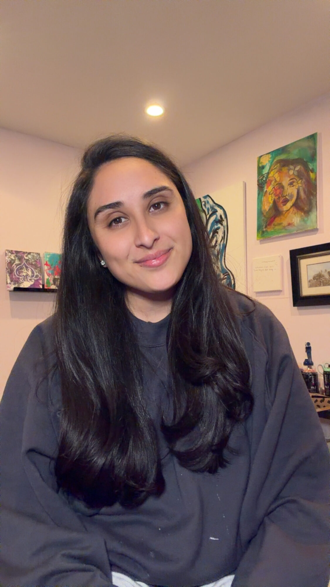 Young woman with long dark hair smiling indoors, paintings on the wall behind her.