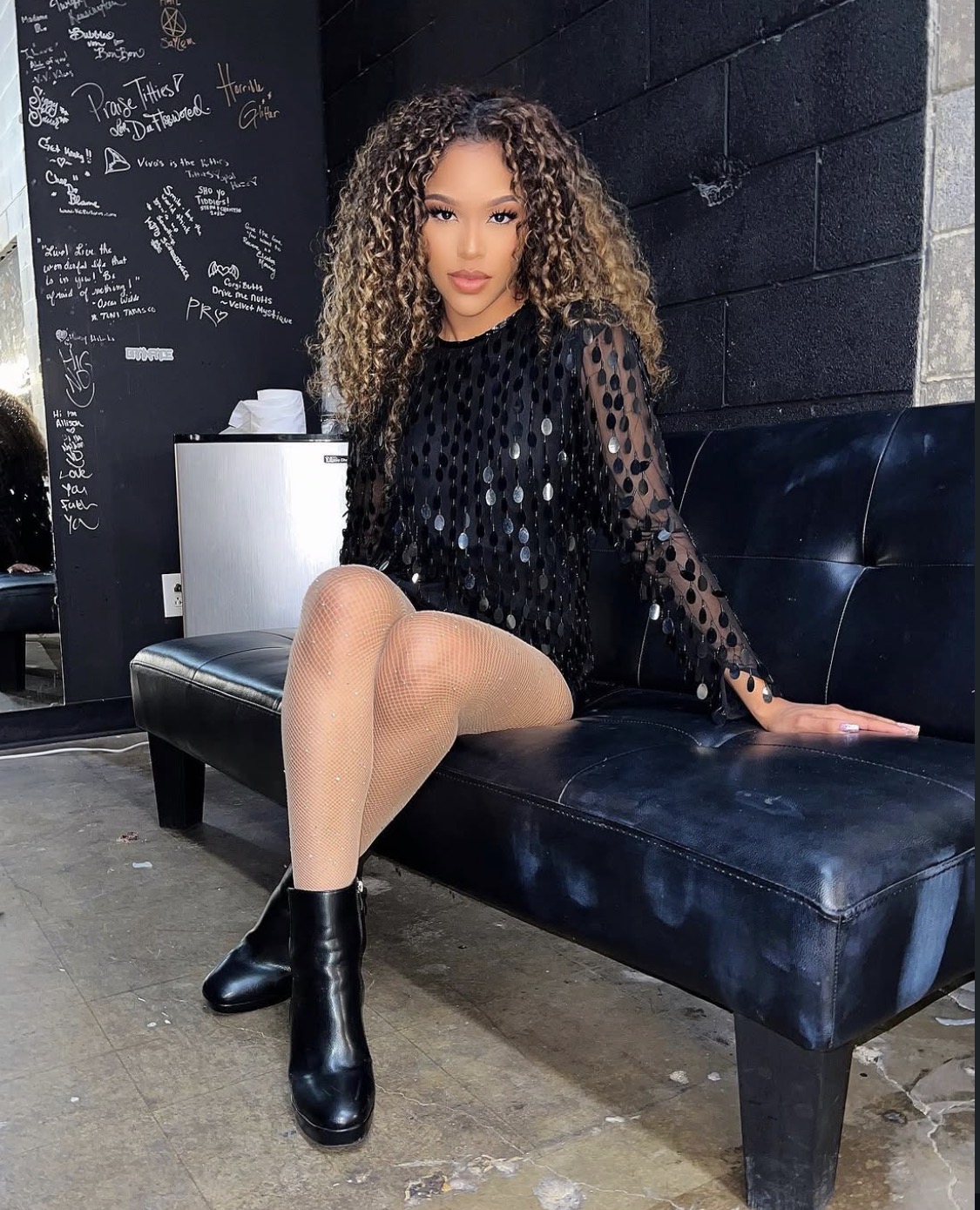 Young woman with curly hair sitting on a black bench in a casual setting, wearing a black dress with sheer polka dots and black boots.
