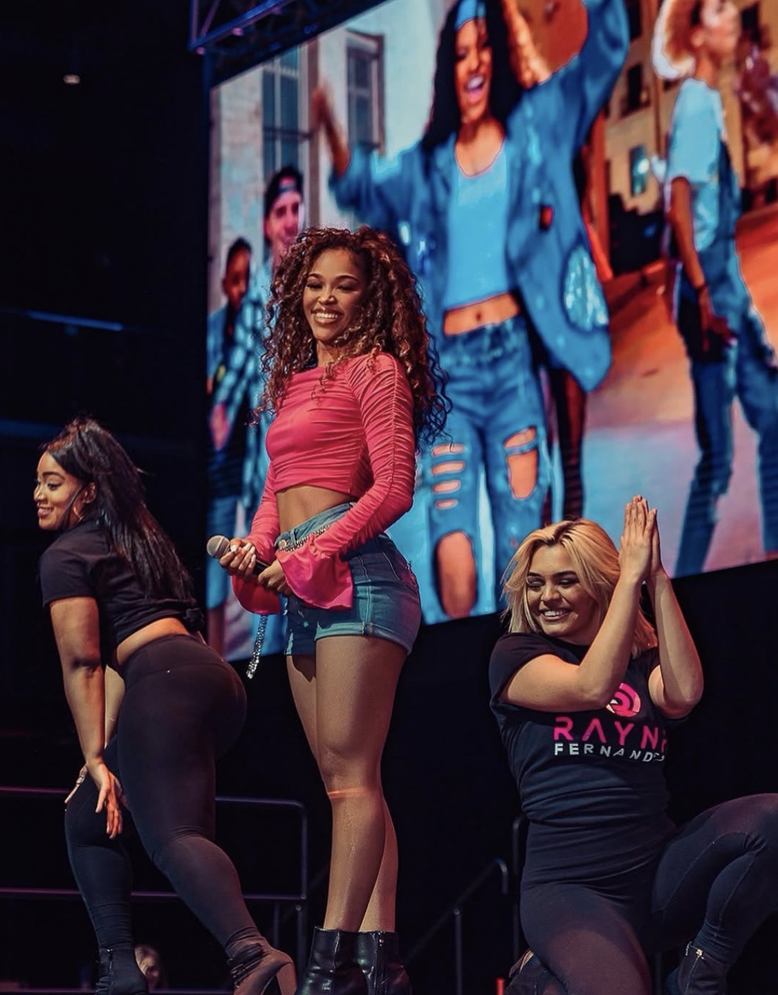 Three women on stage, one smiling and holding a microphone, with a large screen showing a woman in a blue jacket behind them.