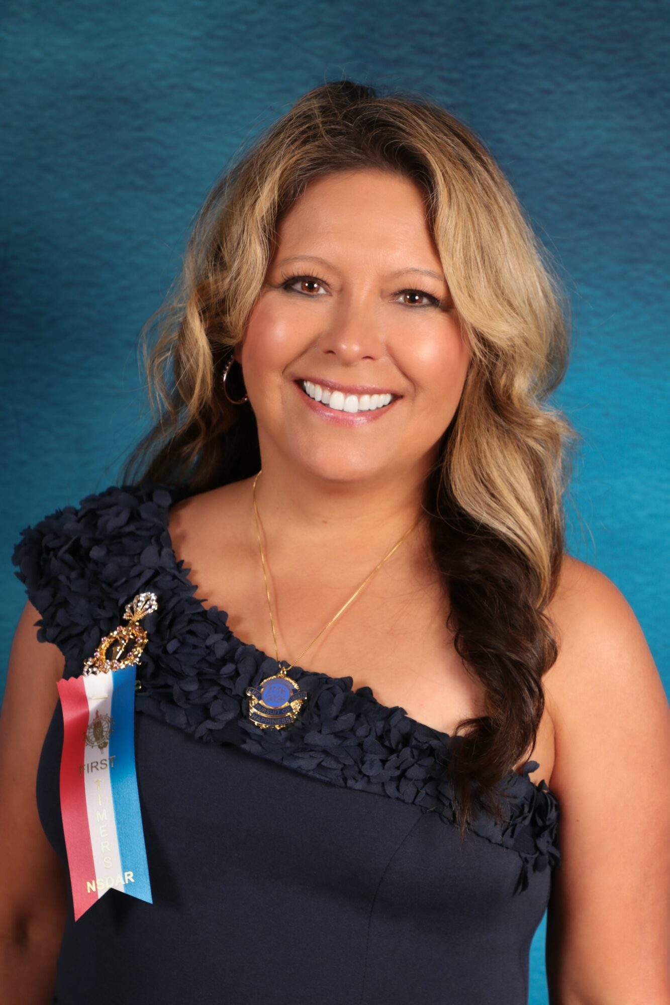 Smiling woman with wavy blonde hair wearing a dark dress and a ribbon with red, white, and blue stripes.