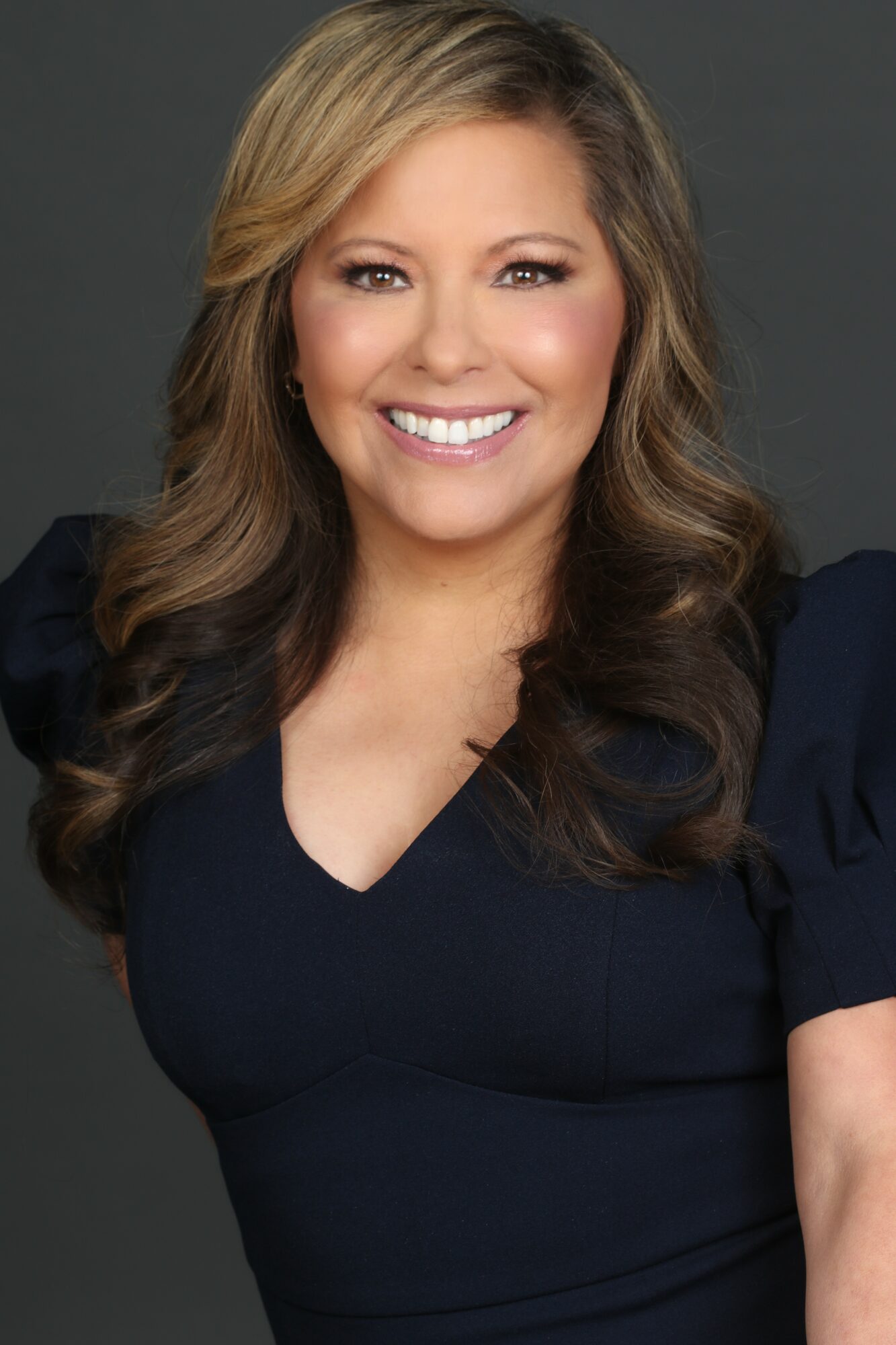 Smiling woman with long wavy hair wearing a dark top against a dark background.