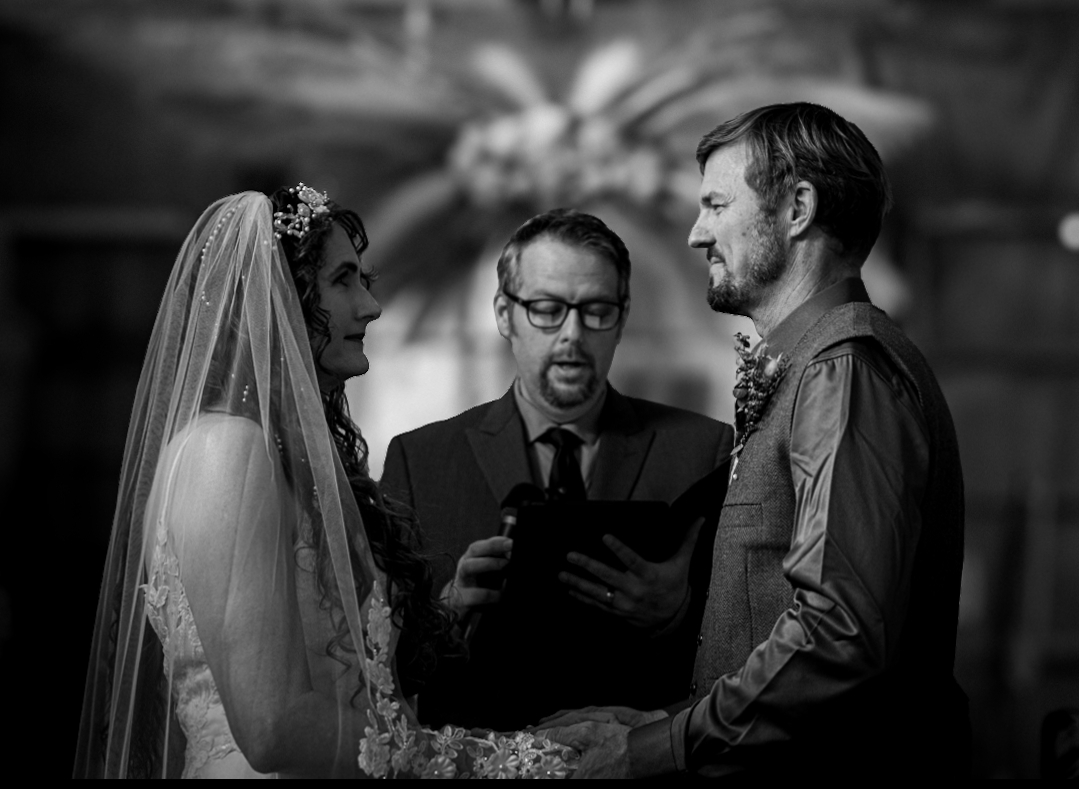 Bride and groom holding hands during wedding ceremony, officiant reading vows, in black and white.