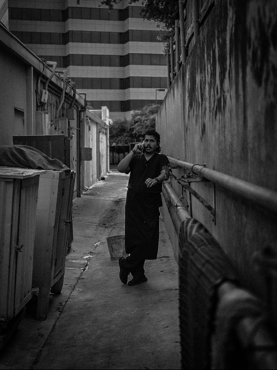 Person walking down a narrow alleyway with buildings on both sides, holding a phone to their ear, in black and white.
