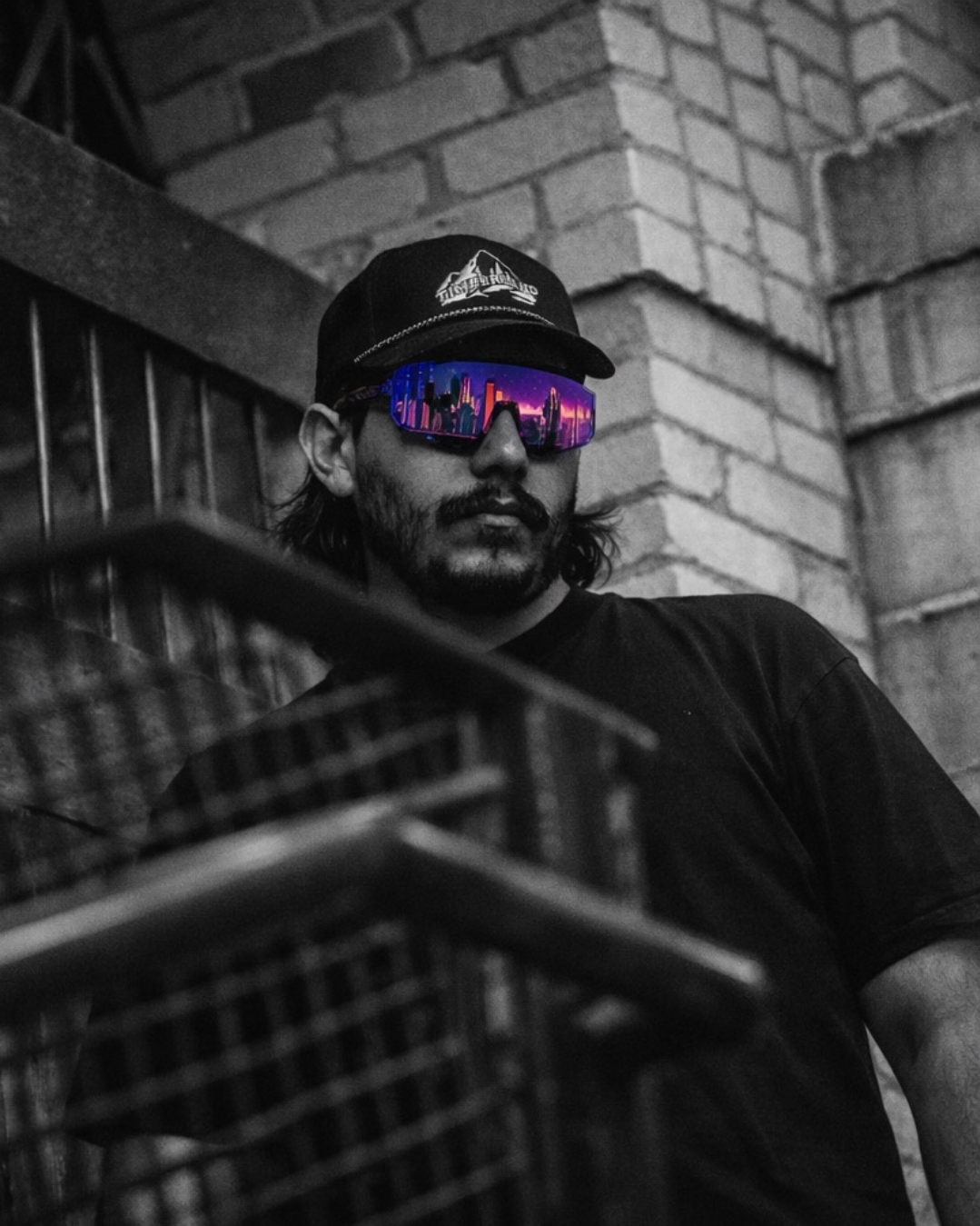Man wearing reflective sunglasses and a cap, leaning against a brick wall, in black and white with colorful reflections in sunglasses.