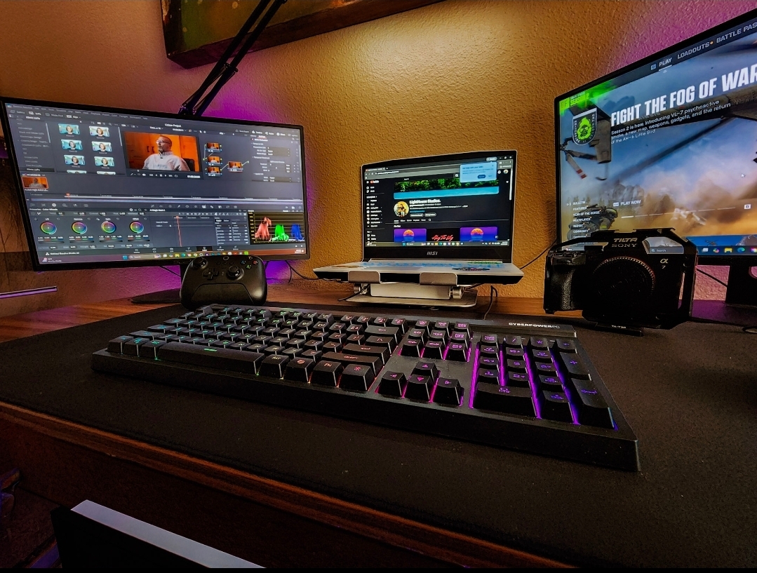 Desk setup with three monitors, a keyboard, and a camera, in a room with orange and purple lighting.