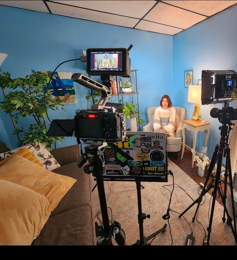 Person sitting on a chair in a blue room with plants, a lamp, and a painting, being filmed with cameras and equipment.