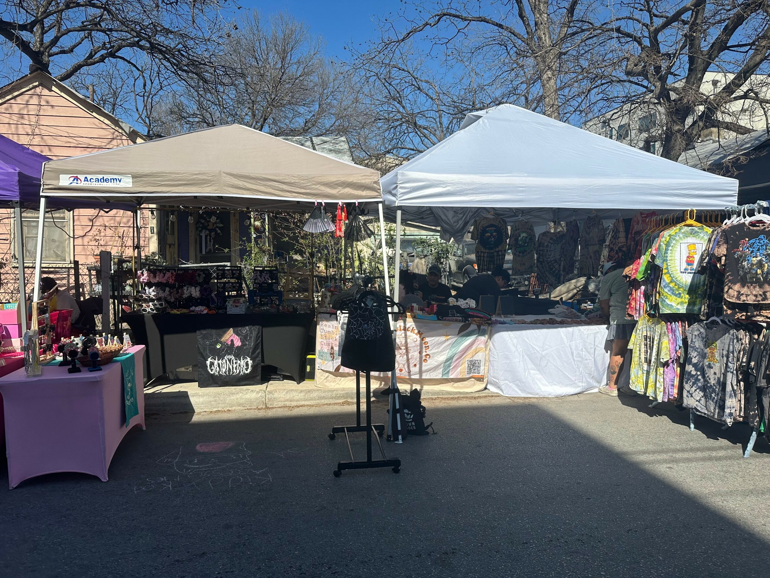 Two tents set up outdoors with tables displaying items, trees and a house in the background, sunny day.