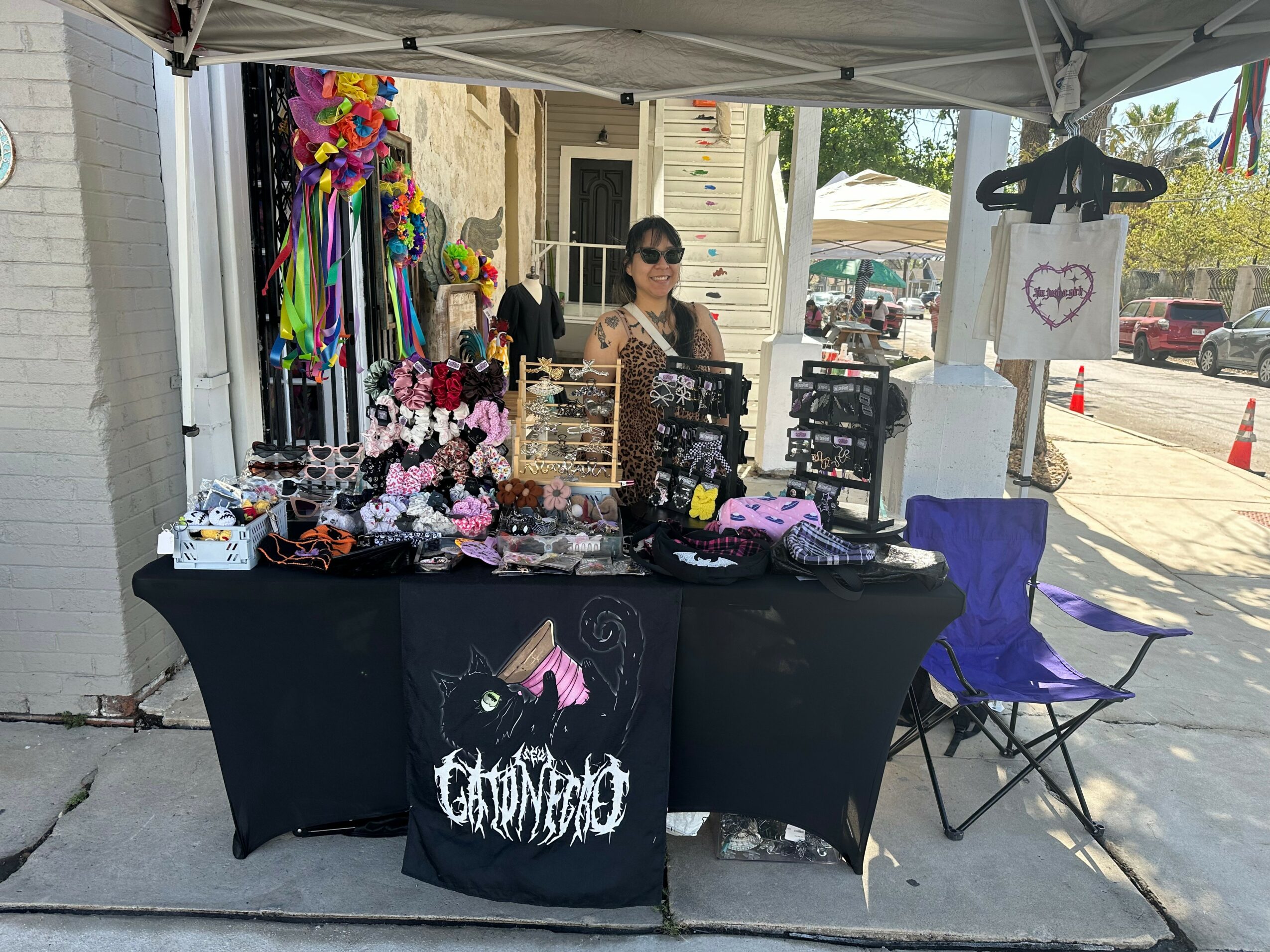 Vendor selling jewelry and accessories at outdoor market stall with a woman behind the table, colorful items displayed, and a T-shirt hanging nearby.