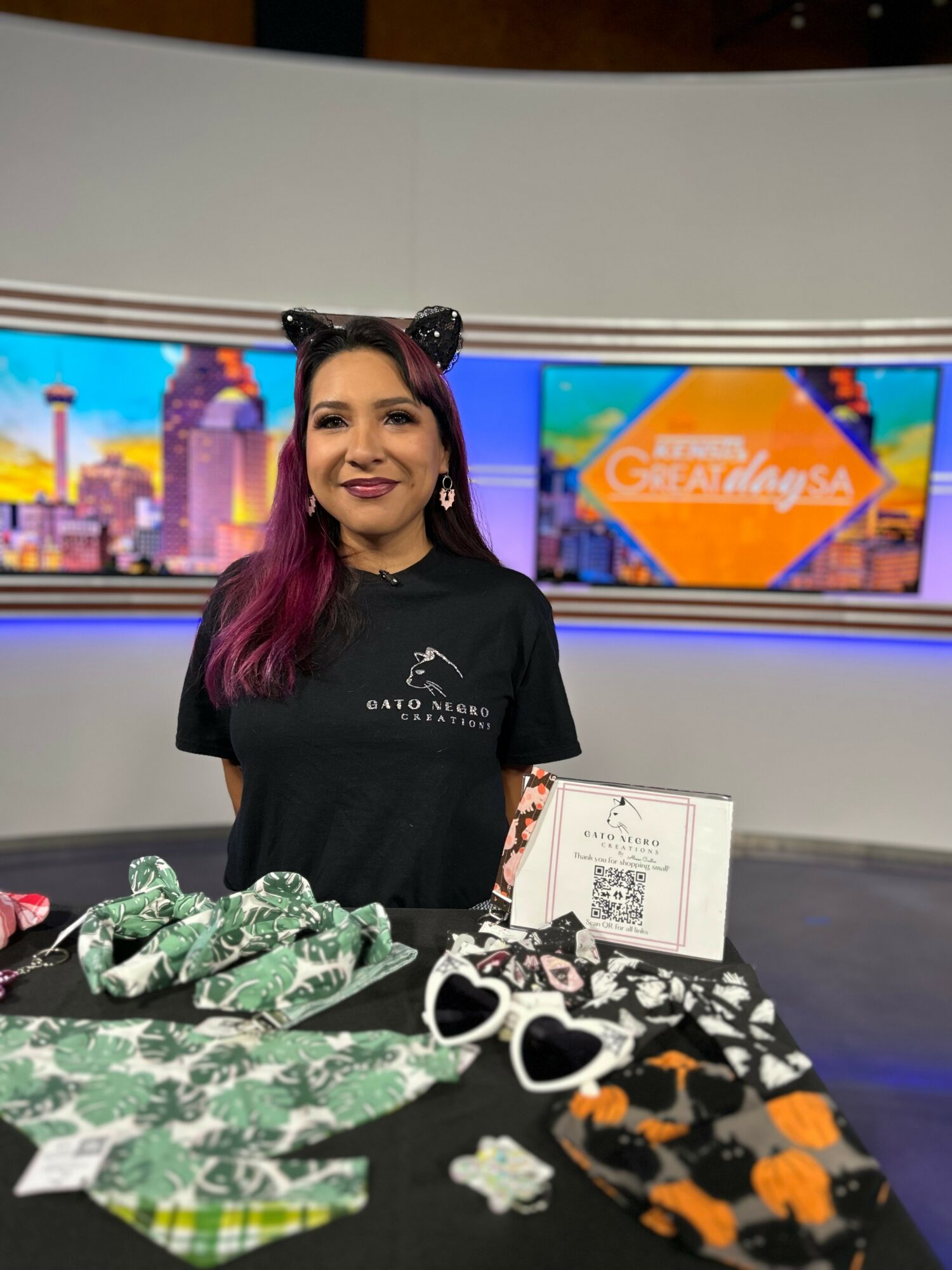 Woman with pink hair smiling behind a table with green, black, and orange face masks, in a studio with cityscape screens.