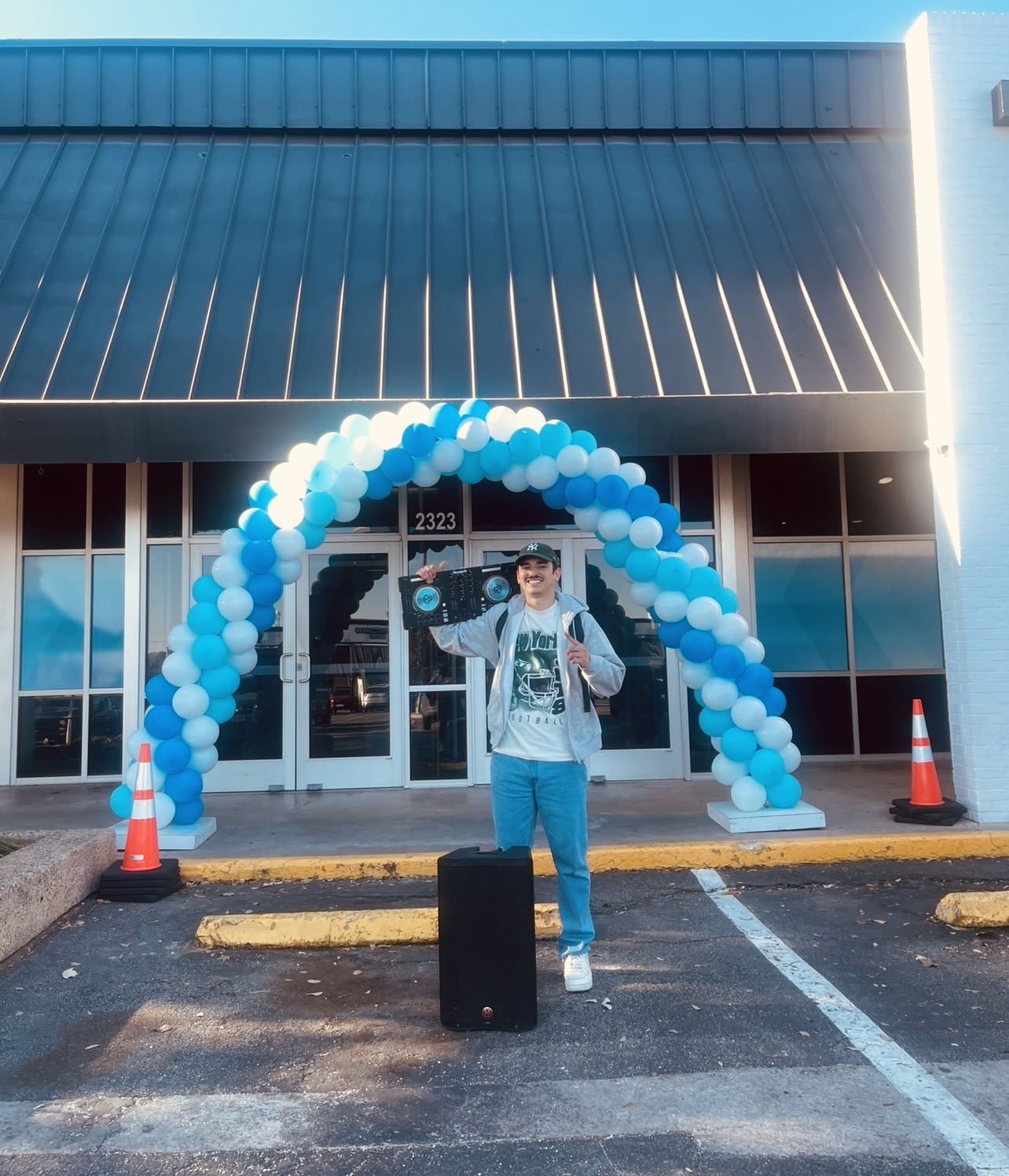 Person standing in front of a building with a balloon arch, holding a suitcase, with parking lot and traffic cones visible.