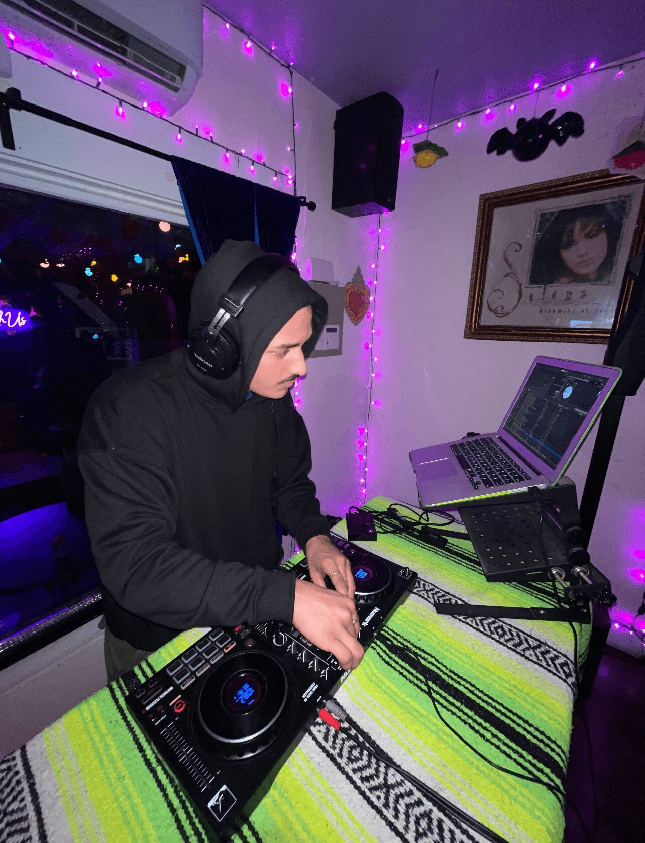Person wearing headphones DJing with equipment in a room decorated with purple string lights and framed picture on wall.