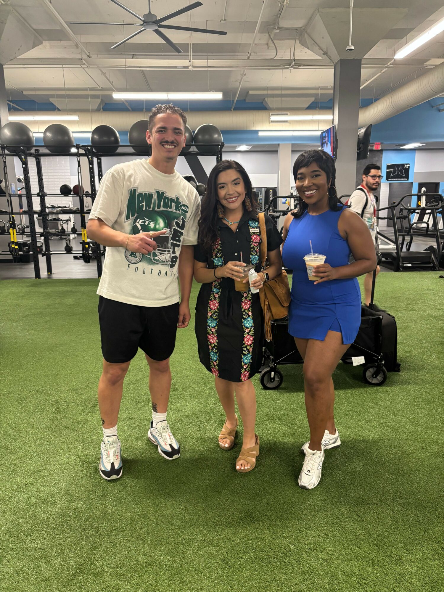 Three women standing on artificial grass in a gym, holding water bottles, smiling, with gym equipment in background.