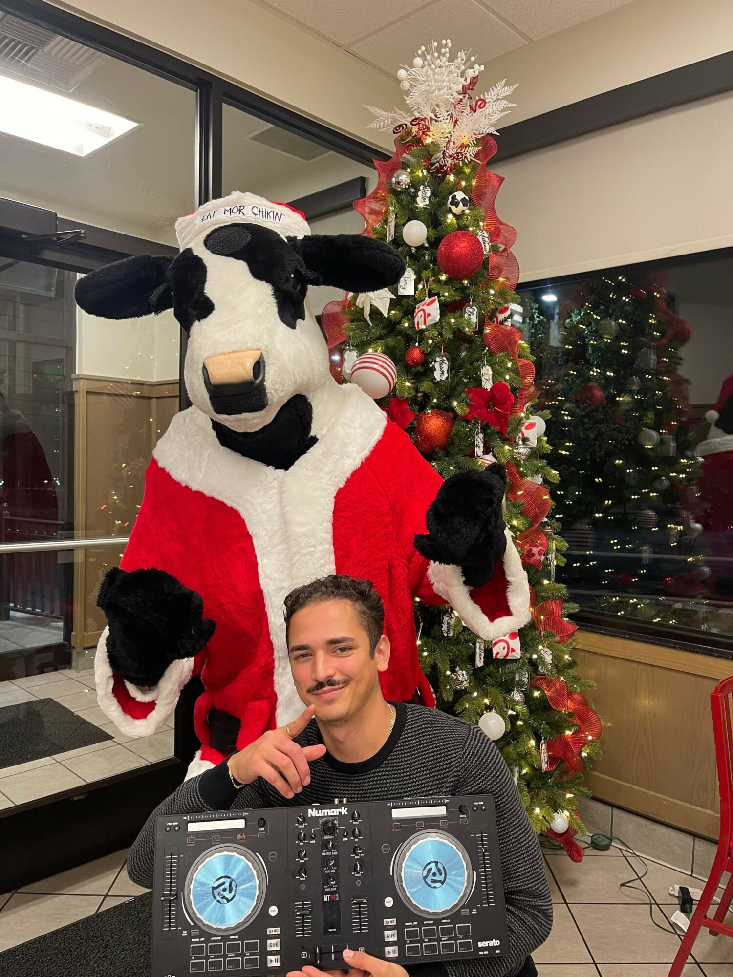Person in Santa costume with a dog, standing in front of Christmas decorations and a DJ controller, smiling.