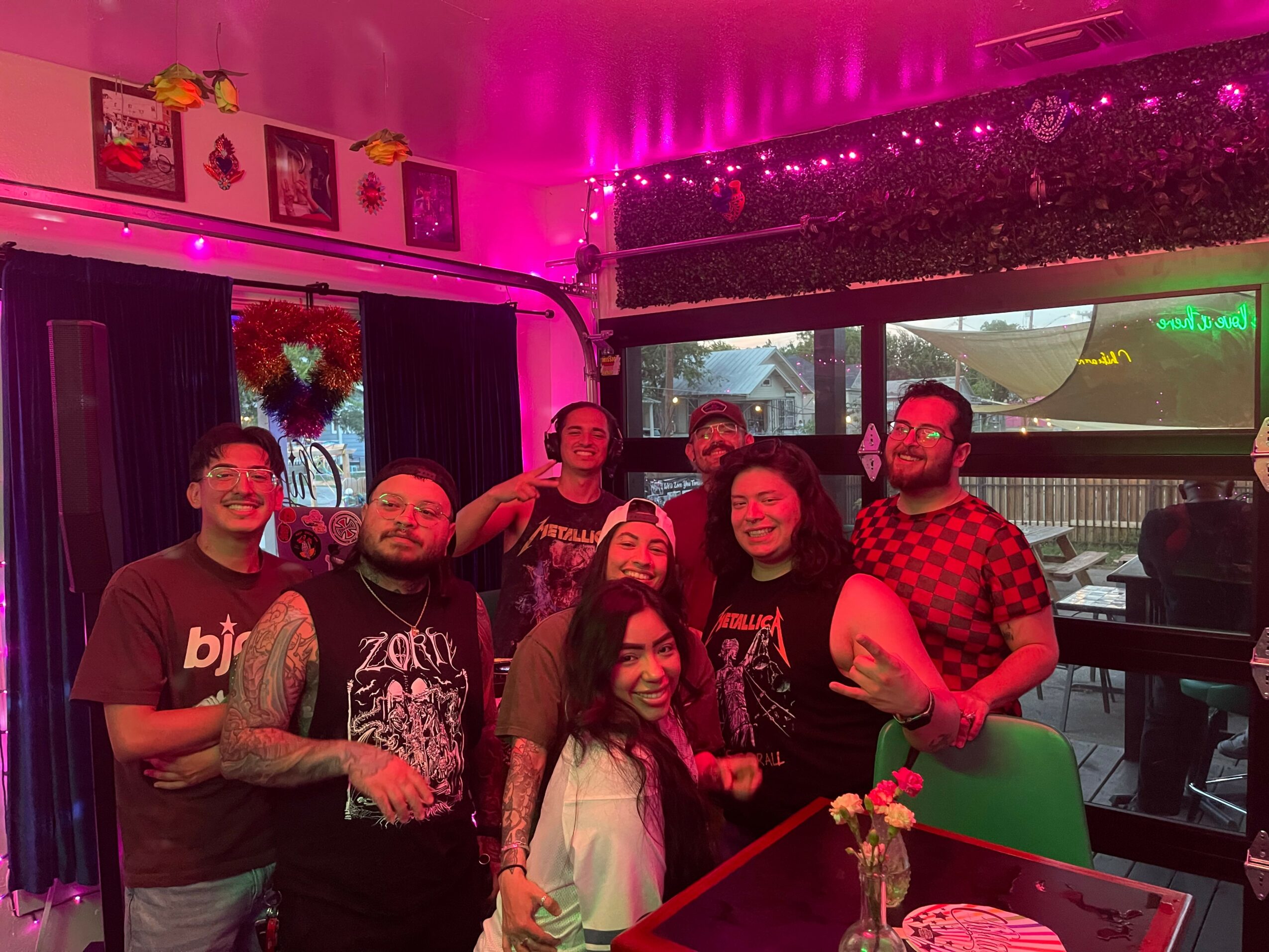 Group of seven people smiling indoors with pink lighting, some holding drinks, in a decorated room with windows and artwork.