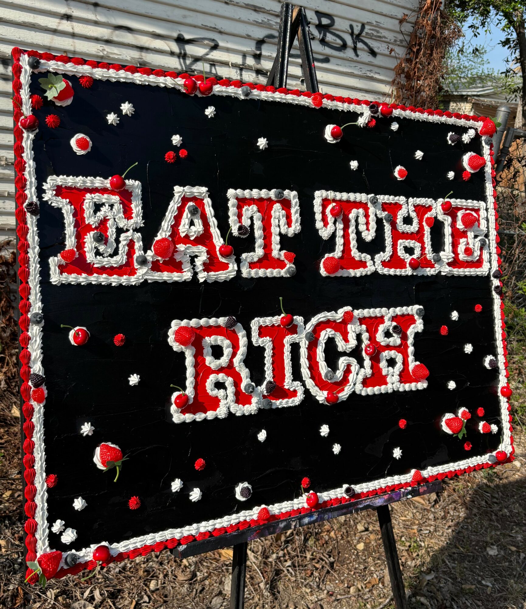 Black sign with red and white decorations reads 'EAT THE RICH' in large letters, decorated with pom-poms and small objects.