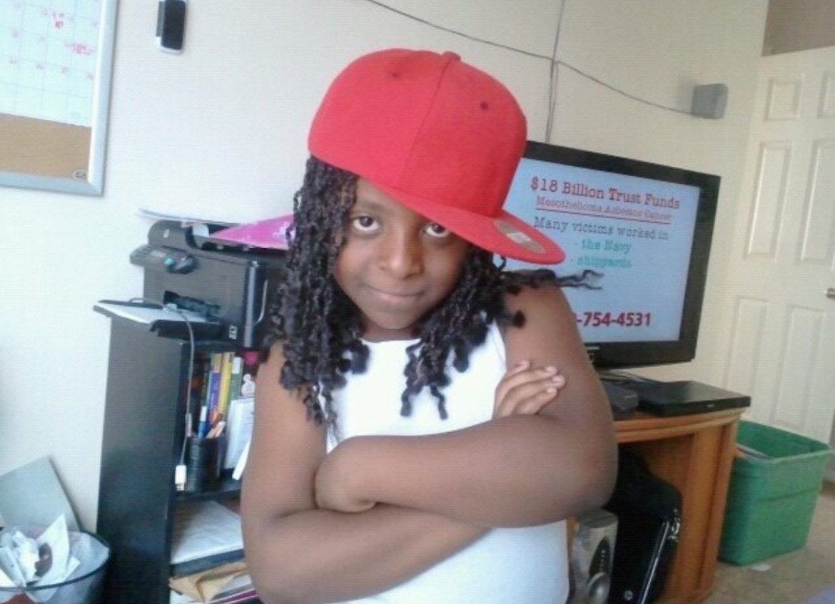 Child with arms crossed wearing a red cap and white shirt, standing in a room with a TV and shelves.