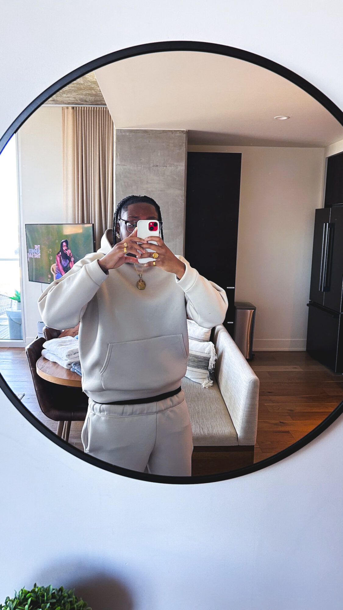 Person taking a mirror selfie in a room with a round mirror, wearing a light-colored outfit, with a sofa and window in the background.