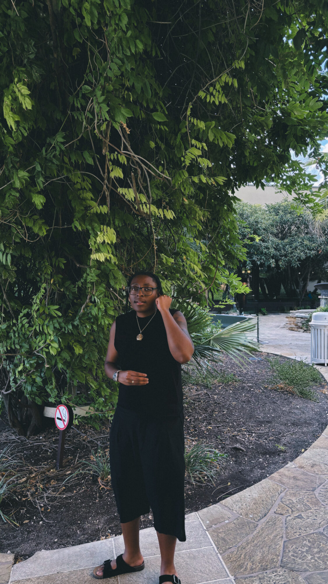 Person standing outdoors near trees, wearing black outfit, holding a phone, with a garden path in background.