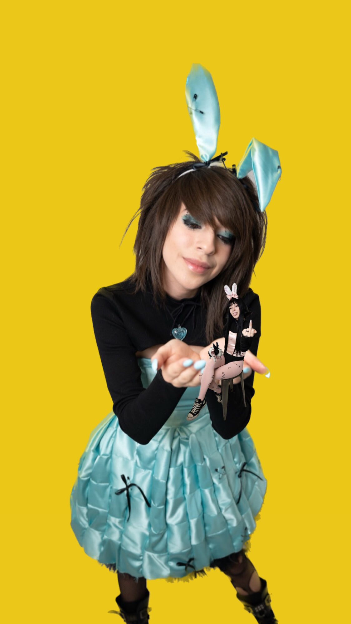 Person with dark hair and makeup wearing a black top and a blue patterned skirt, holding a small object, with blue paper decorations on head, against yellow background.