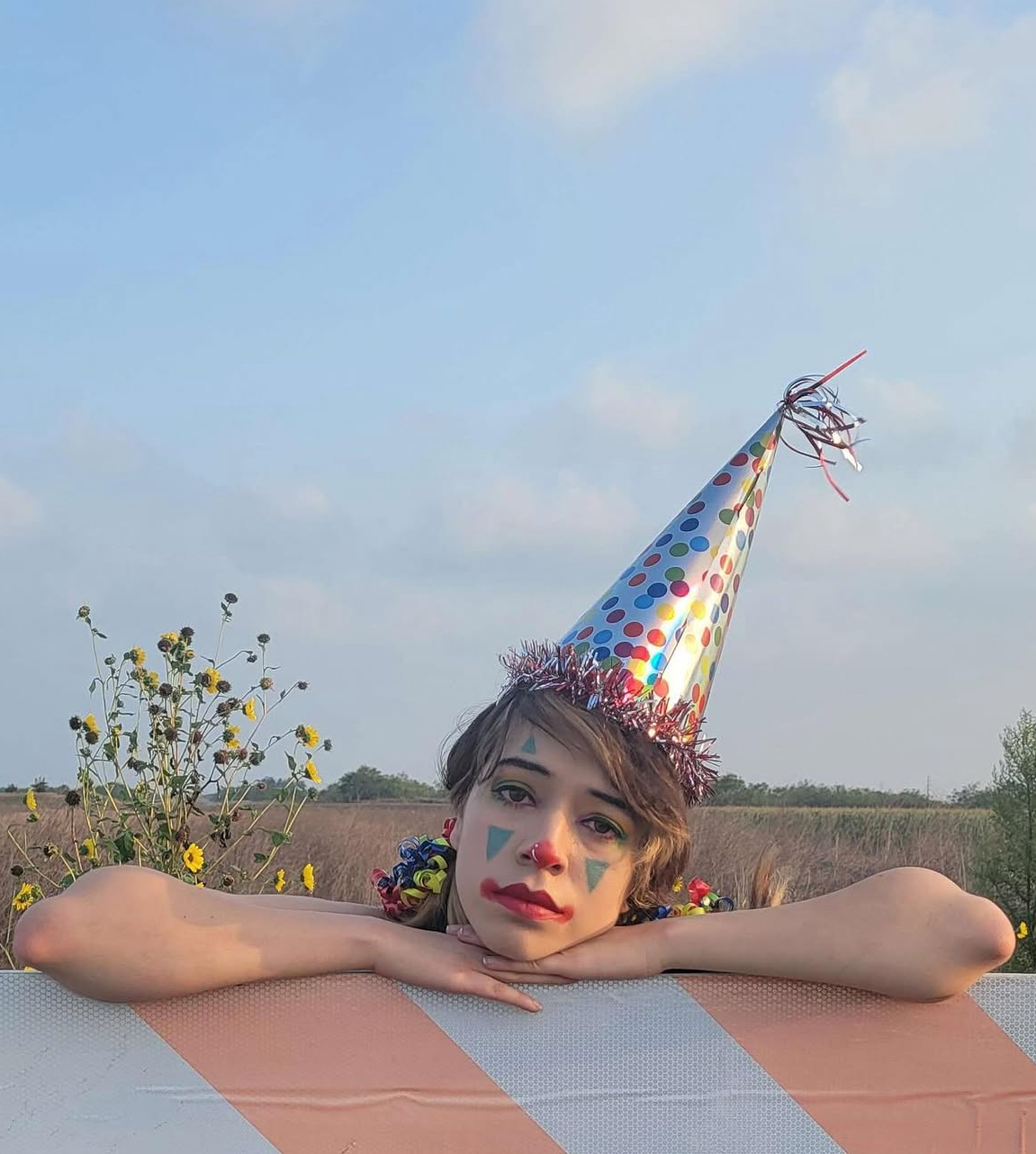 Child with face painted, wearing a colorful party hat, resting chin on arms outdoors with a field and sky background.