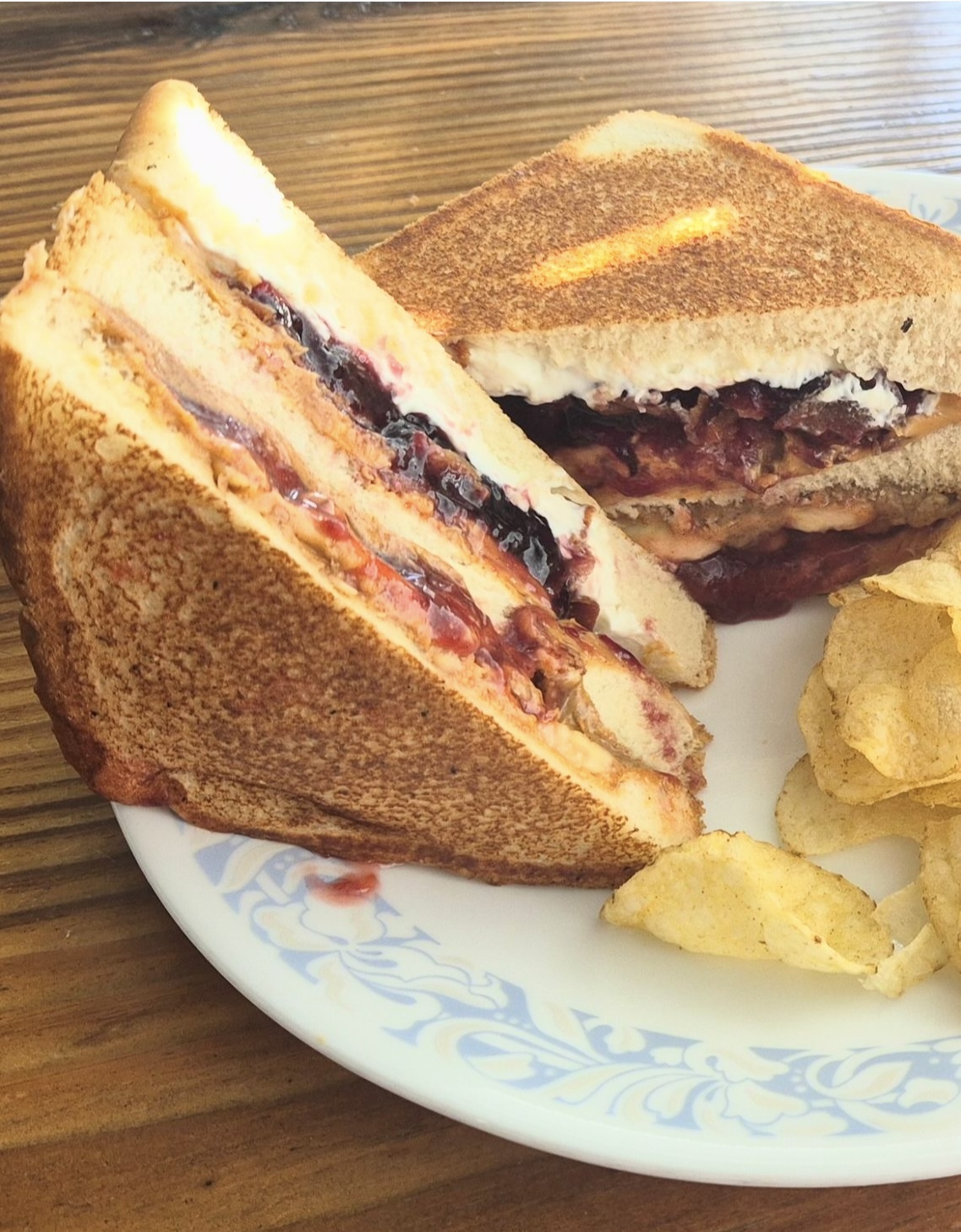 Two toasted sandwiches with layers of meat and cheese, cut in half, on a white plate with potato chips, on a wooden table.