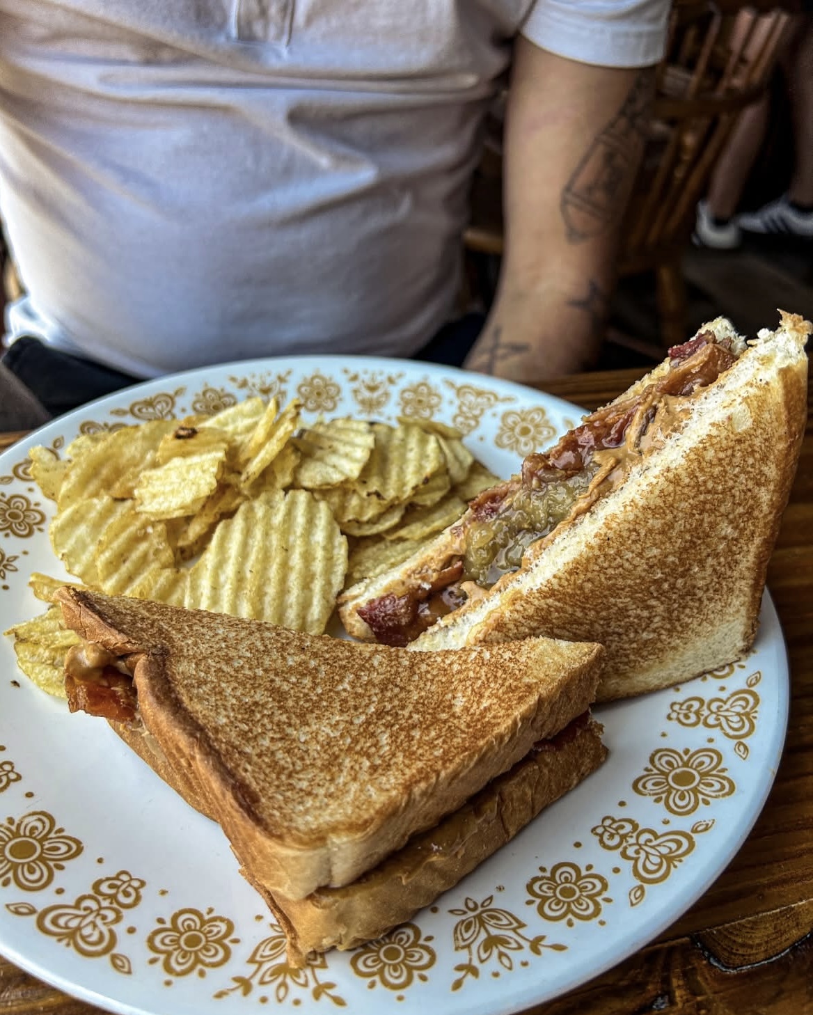 A plate with a toasted sandwich and ridged potato chips on a table, with a person wearing a white shirt in the background.