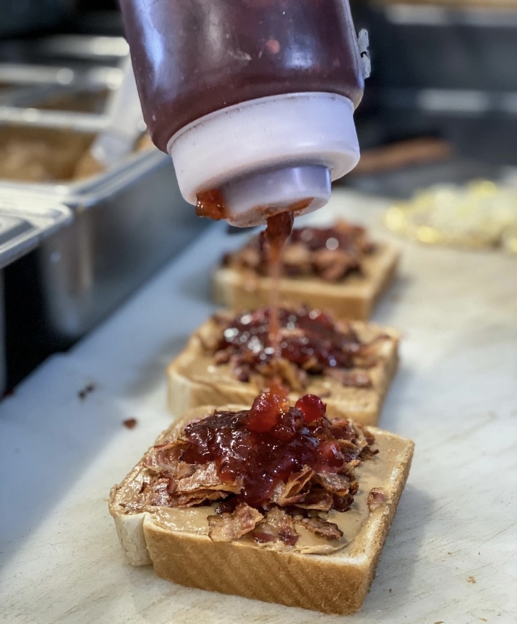 Squeeze bottle dispensing sauce onto open-faced sandwiches on a marble surface, with toppings and kitchen background.