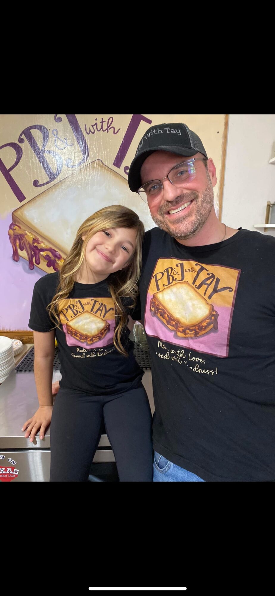 Two people smiling, wearing matching PBJ TAYLOR shirts, standing in front of a decorated wall with a large PBJ sandwich illustration.