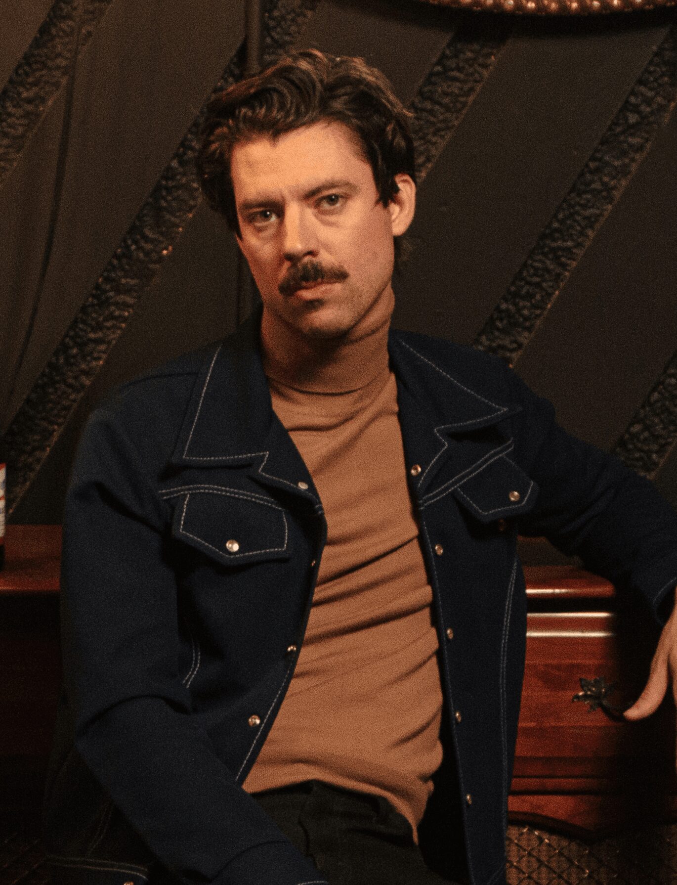 Man with dark hair and mustache wearing a brown shirt and dark jacket, sitting indoors.