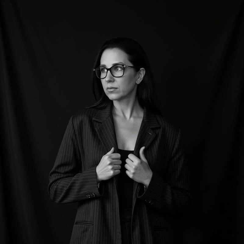 Woman with glasses and dark hair, wearing a pinstripe blazer, looking to the side, holding the lapels, against a black background.